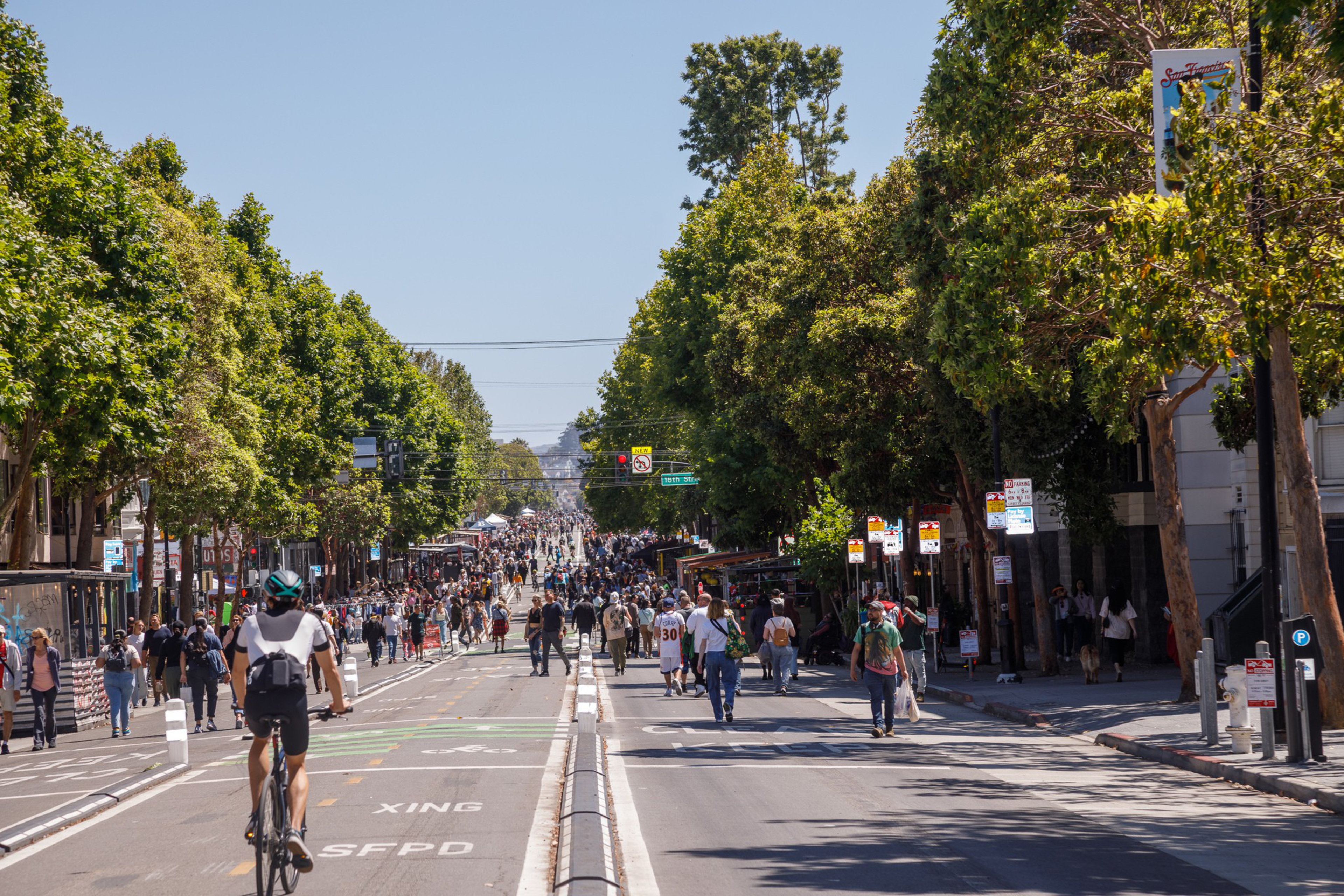 A bustling city street lined with trees, filled with pedestrians and a cyclist, under a clear blue sky.