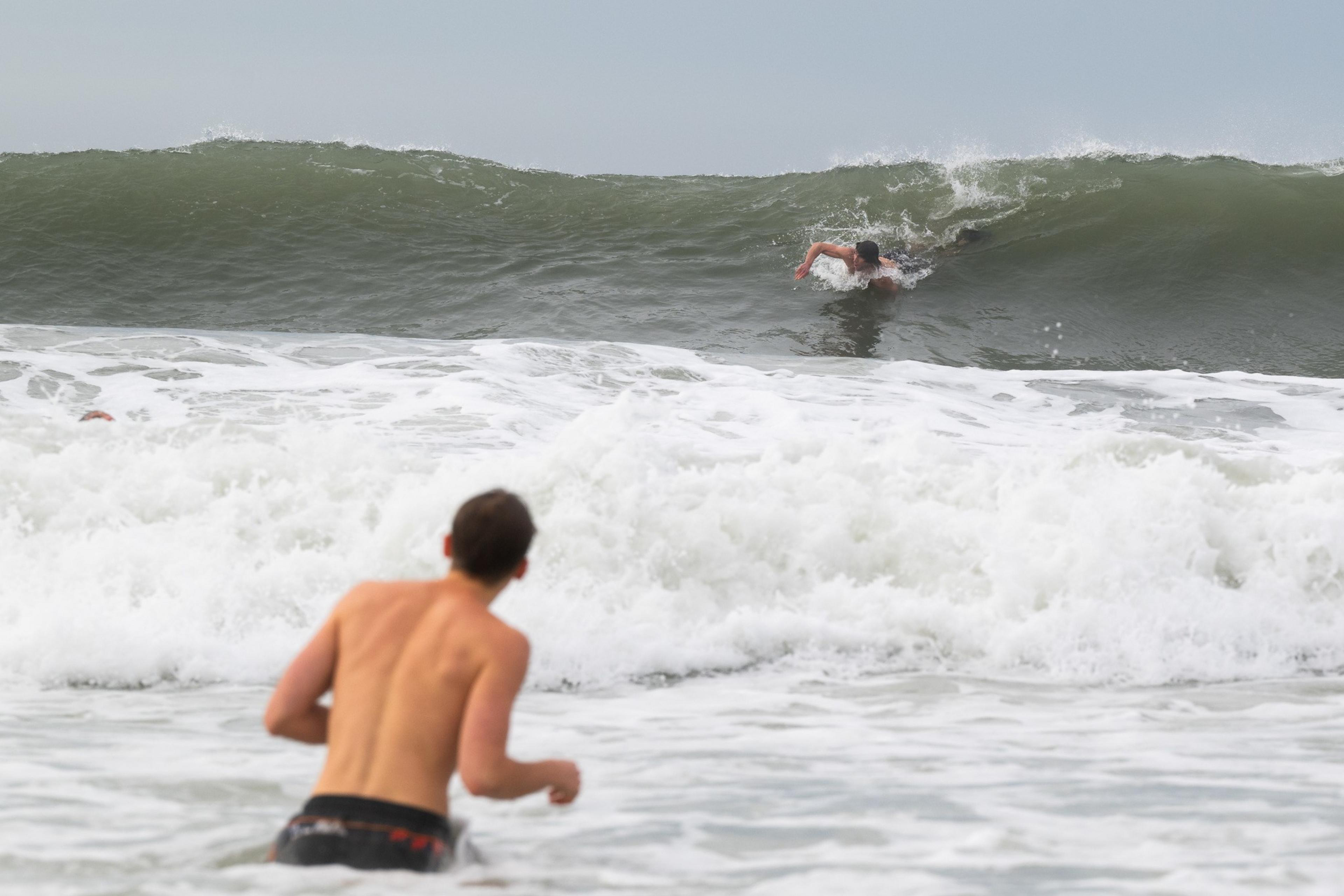 A person is body surfing on a large wave, while another stands in the foreground observing. The ocean is frothy and active under a cloudy sky.