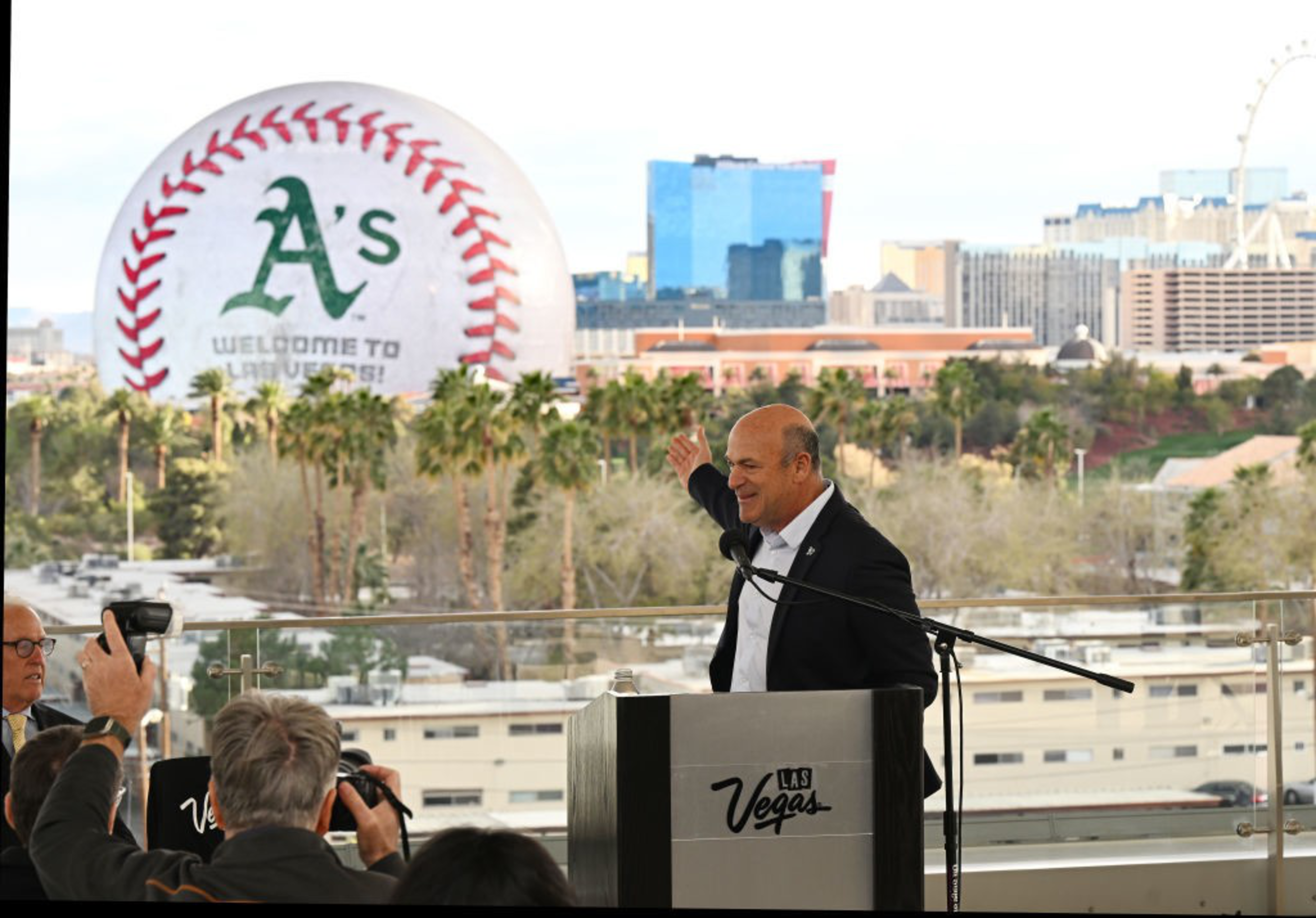 A man stands at a podium labeled 'Las Vegas,' addressing an audience. Behind him, a giant baseball with &quot;A's Welcome to Las Vegas!&quot; is visible, set against a cityscape.