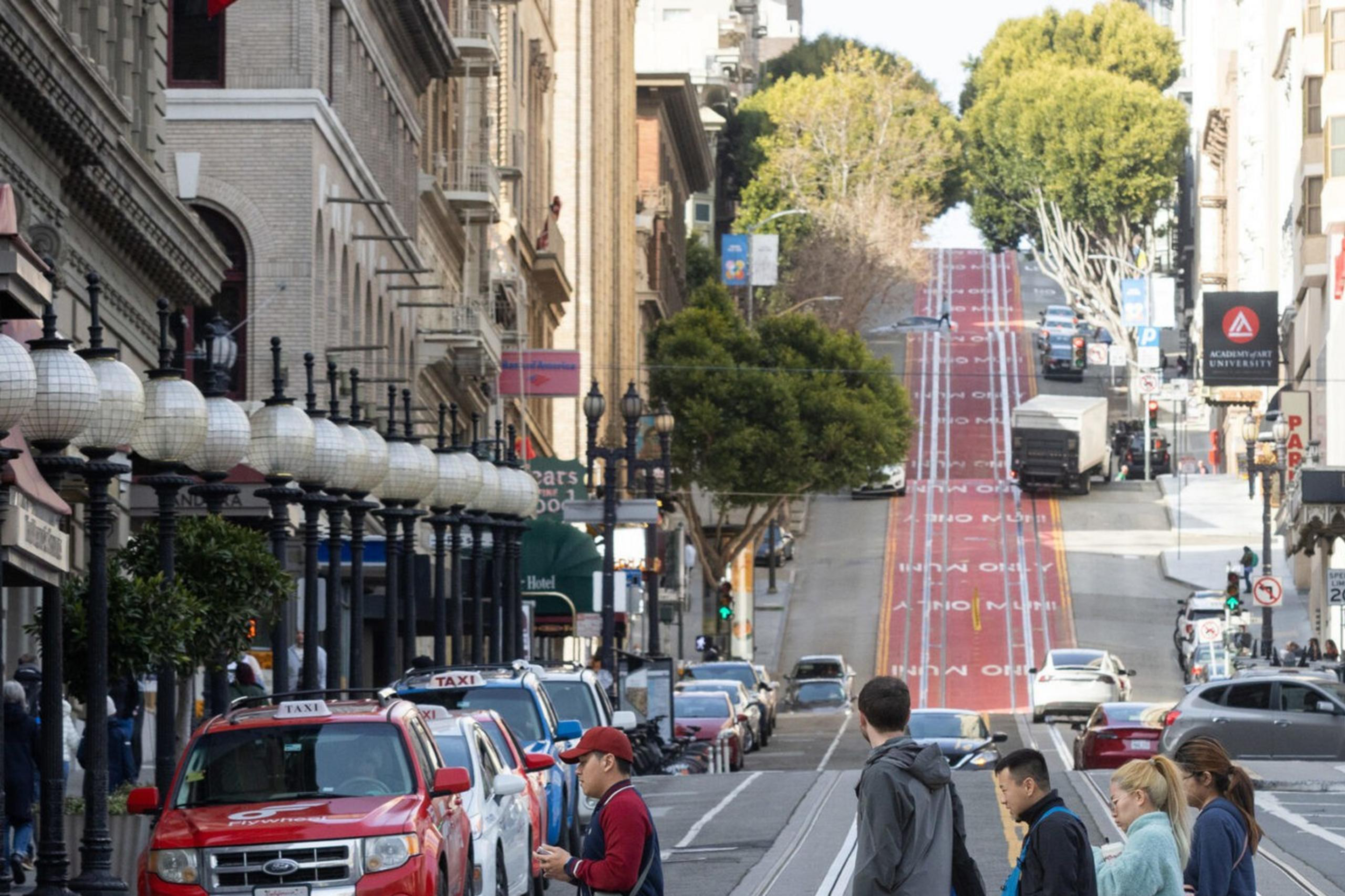 The image shows a bustling city street with a steep hill in the background. People are crossing the street, cars and taxis are lined up, and a bus is on the right side.