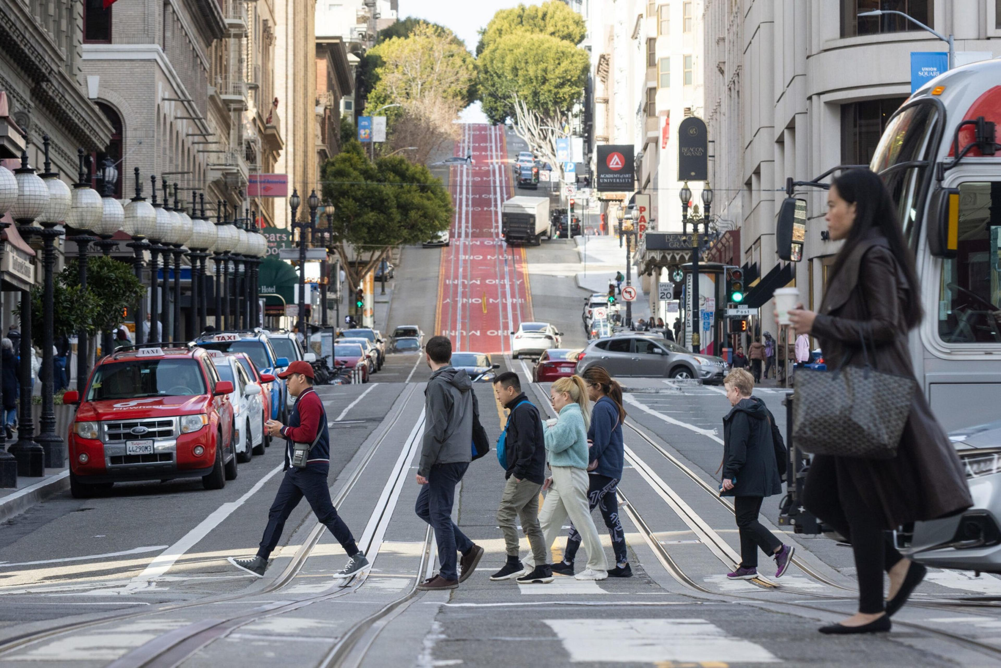 The image shows a bustling city street with a steep hill in the background. People are crossing the street, cars and taxis are lined up, and a bus is on the right side.