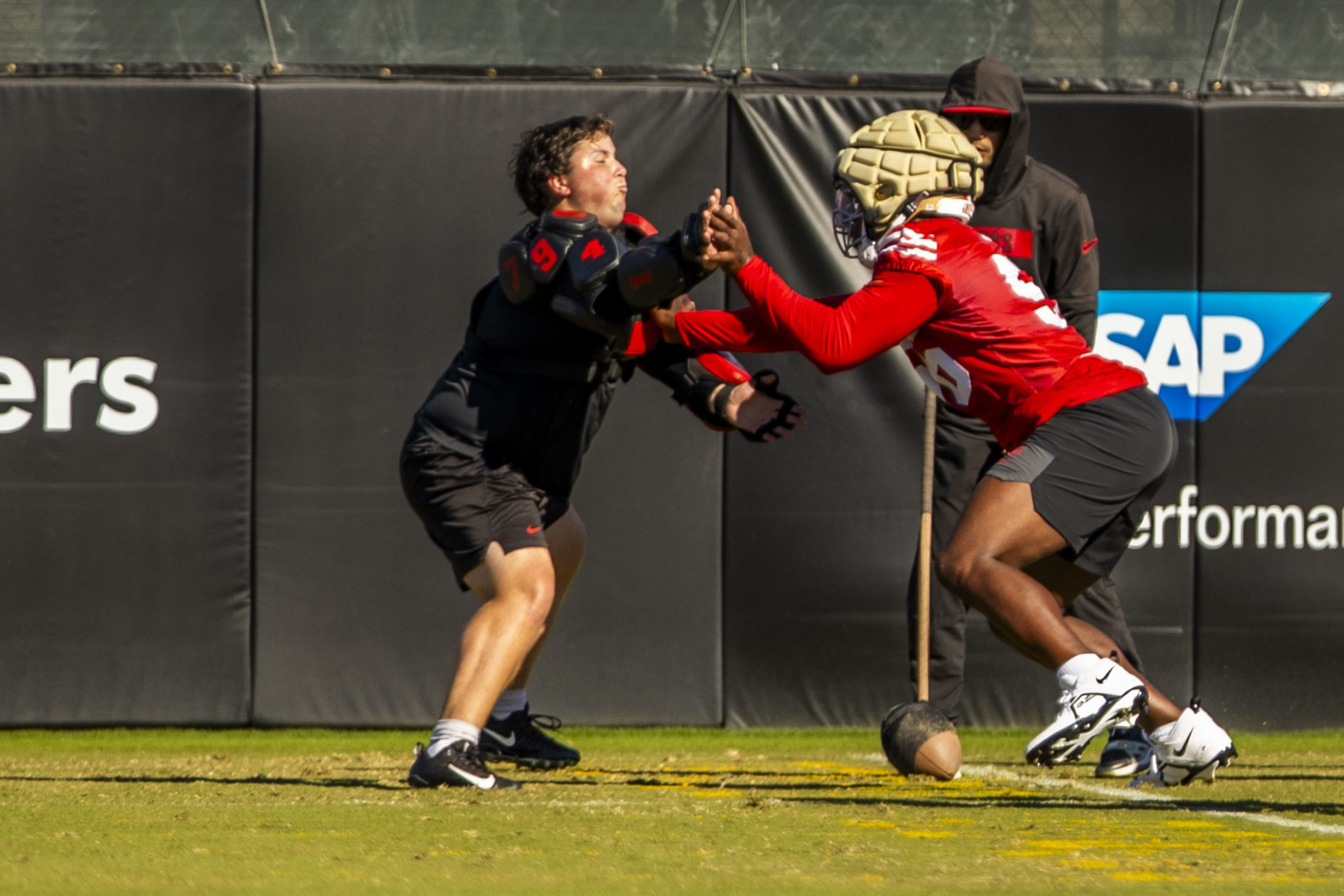 Two football players, one in black and one in red with a padded helmet, are engaged in a blocking drill on a grassy field with a coach observing nearby.