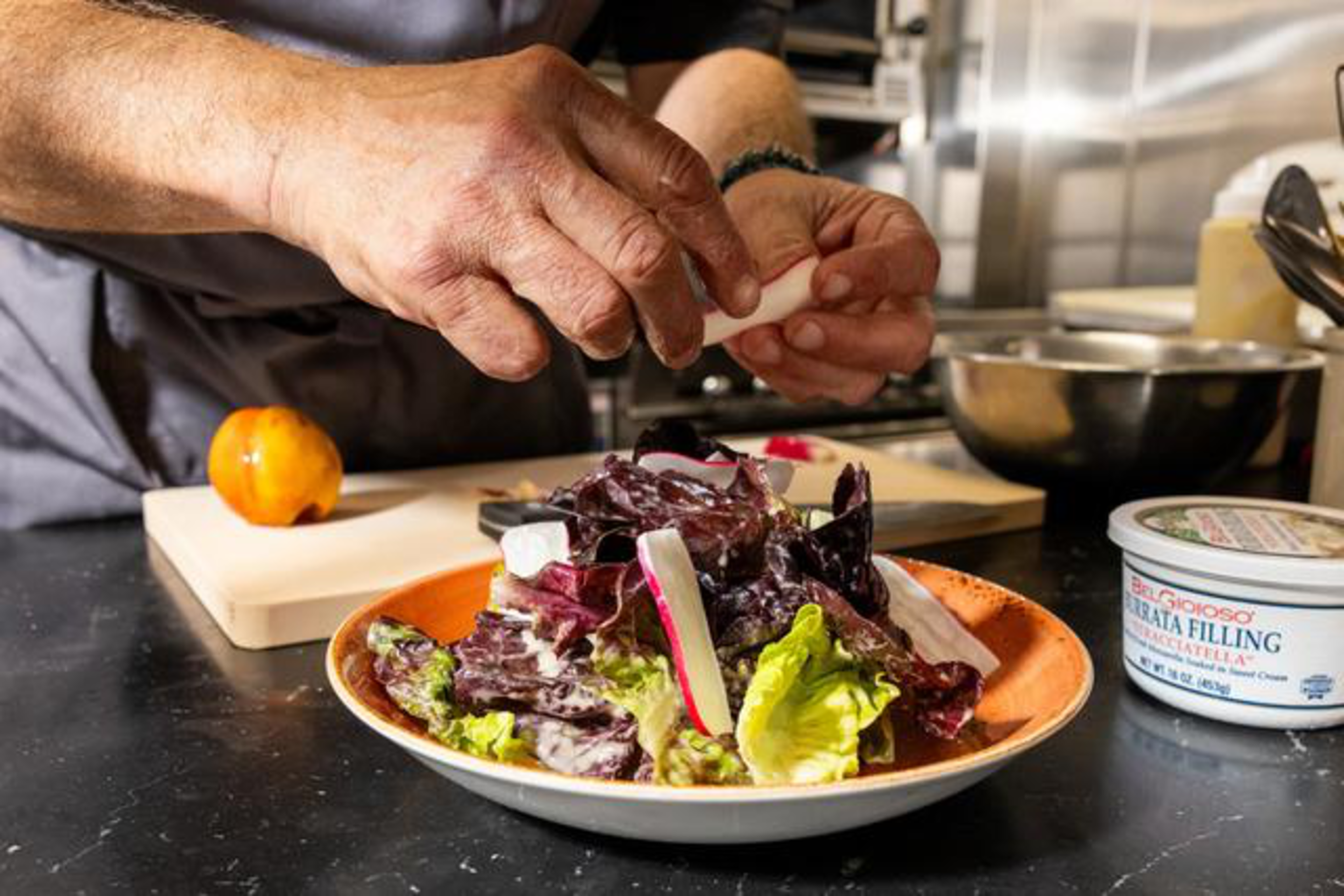 Hands are placing sliced radishes on a fresh salad with mixed greens on a plate, with a cutting board, a tomato, and a container of burrata filling nearby.