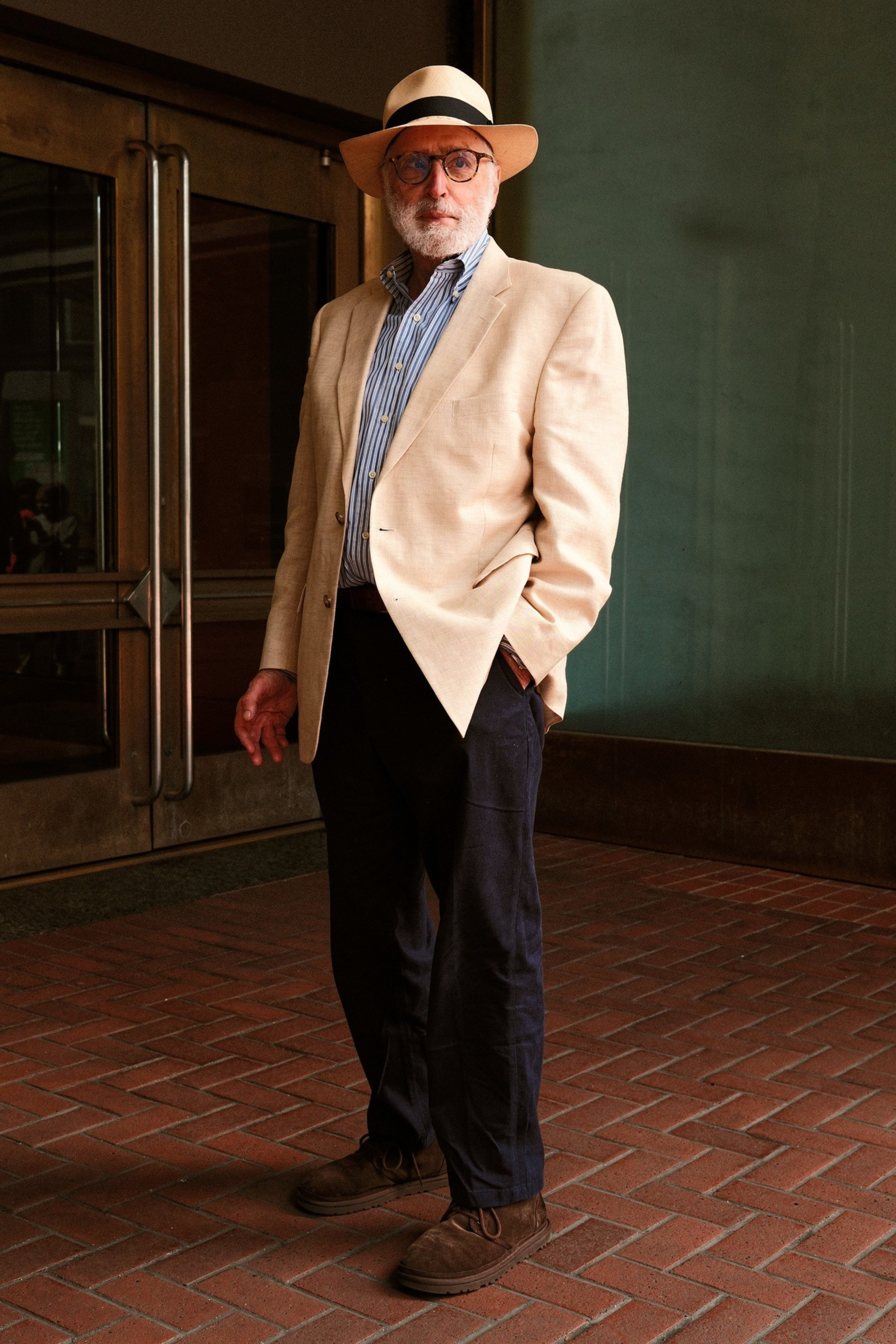 An older man with a white beard wears a beige blazer, striped shirt, dark pants, brown shoes, round glasses, and a beige hat with a black band.