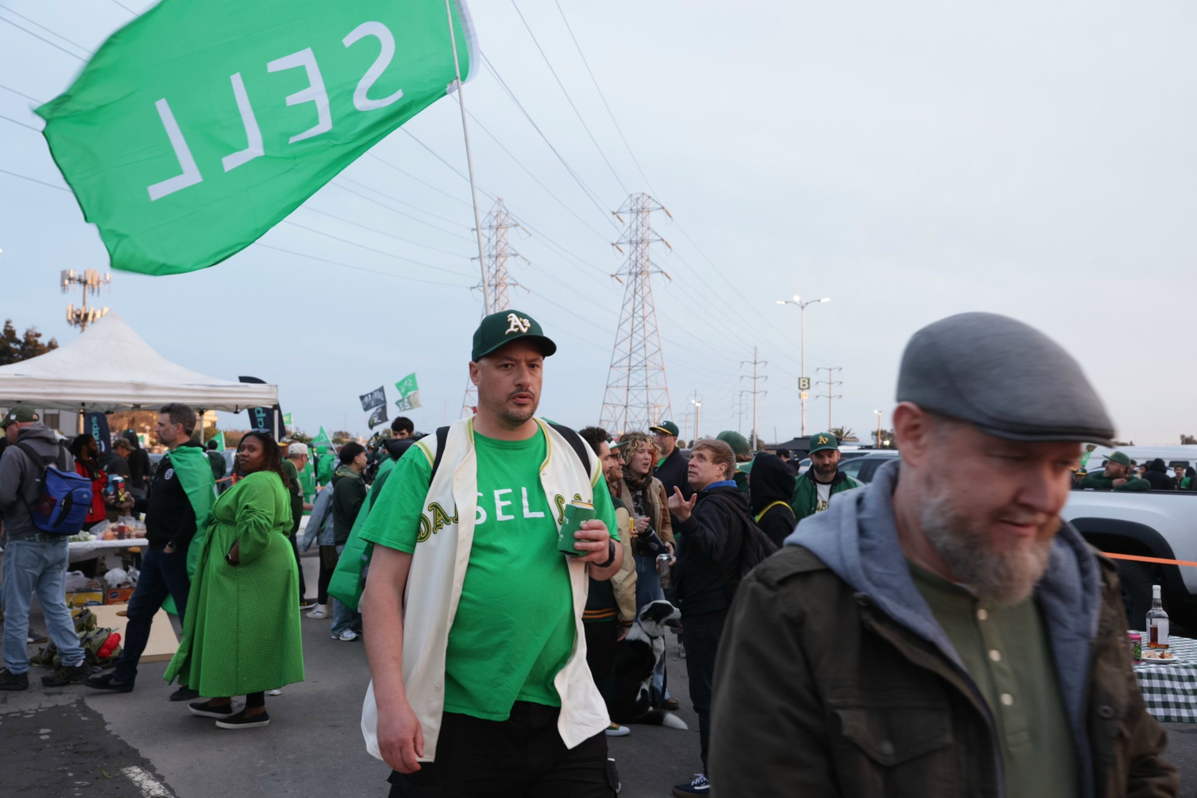 A crowded outdoor gathering with people in green attire, some with Oakland A's logos, and a SELLS flag.