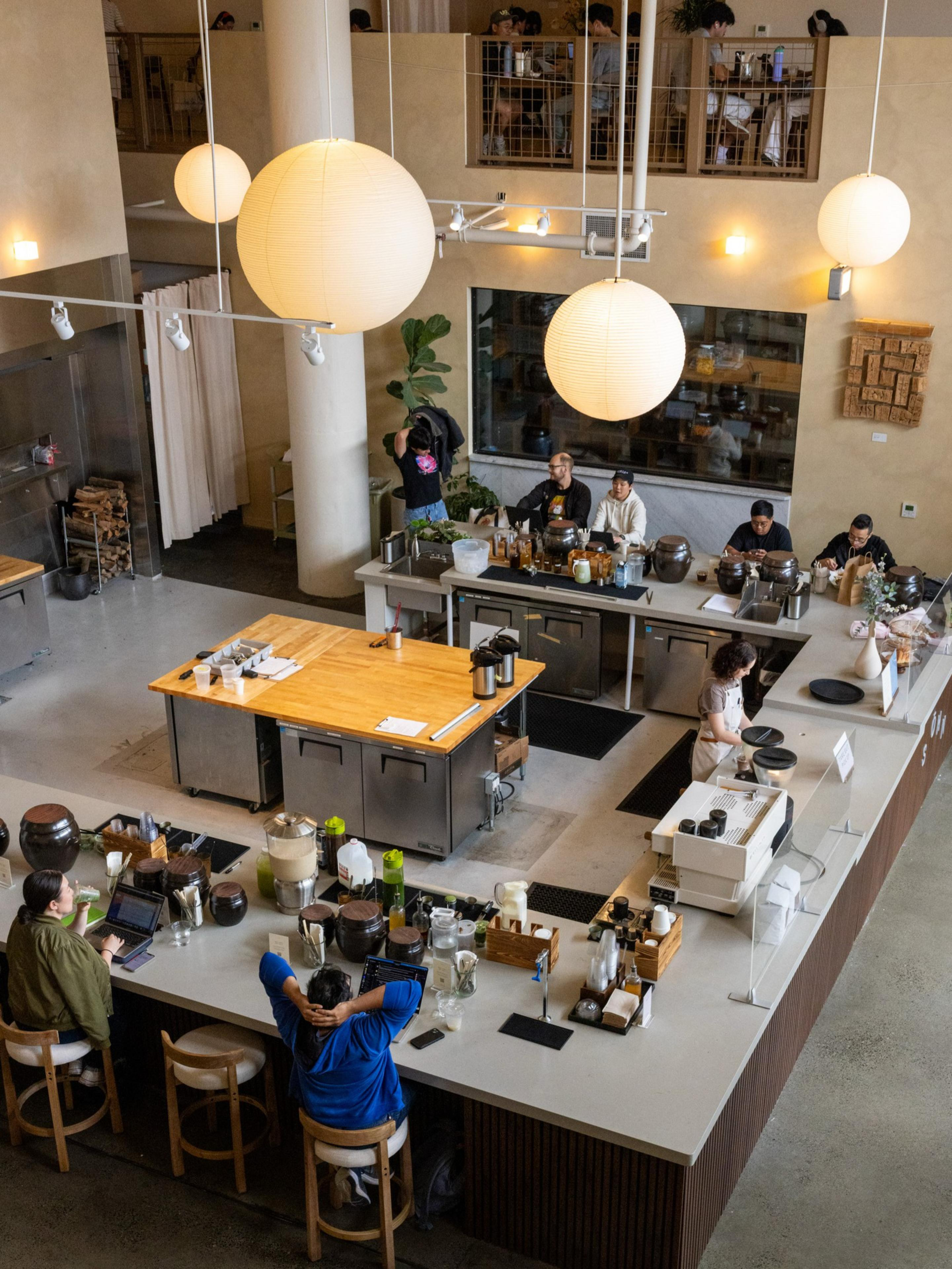 A modern coffee shop with a large central counter, people sitting and working, and hanging round paper lanterns illuminating the space.