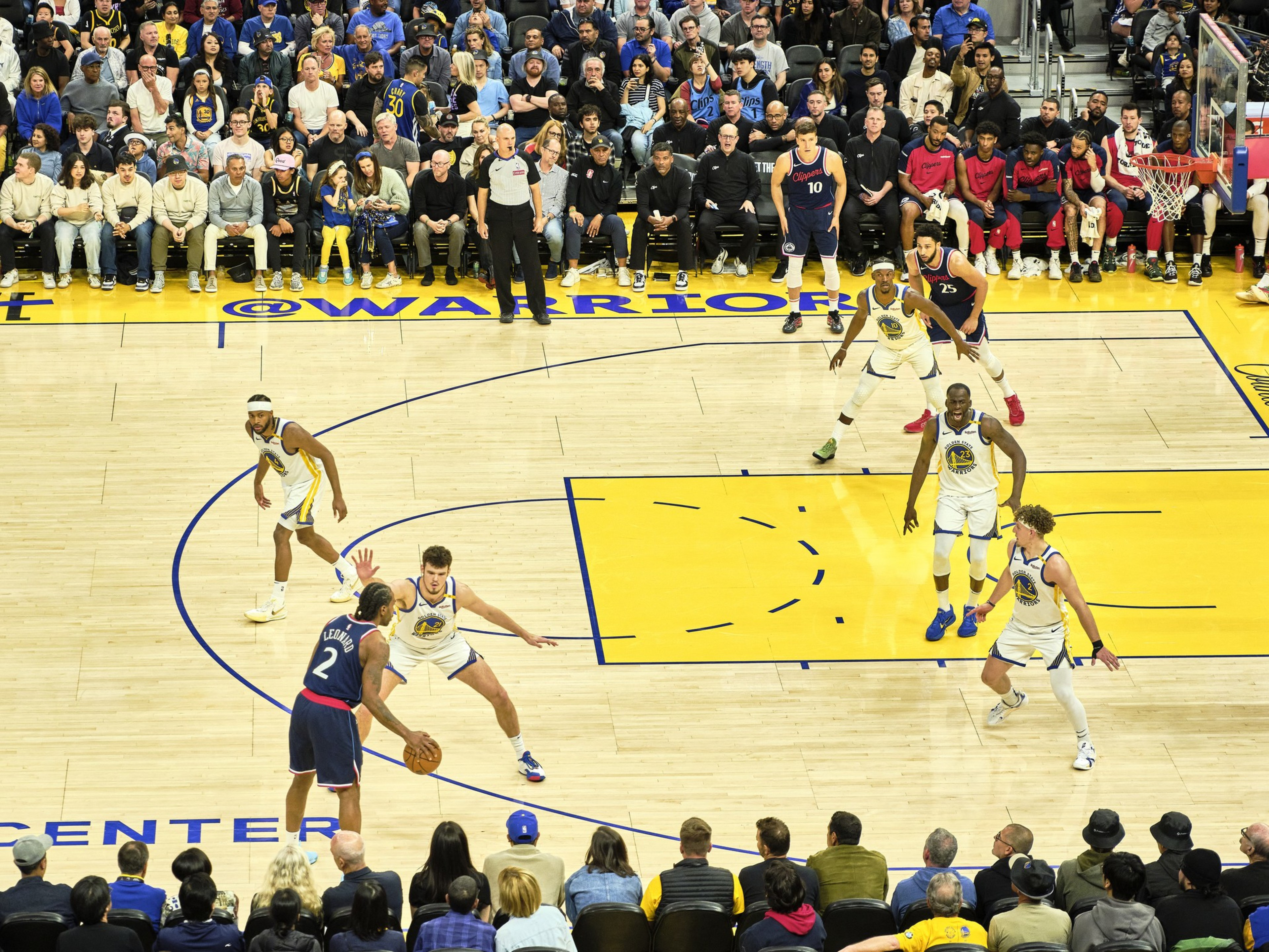 A basketball game is underway with players in blue and white uniforms in action. The court is surrounded by a packed audience, and the game is intense.