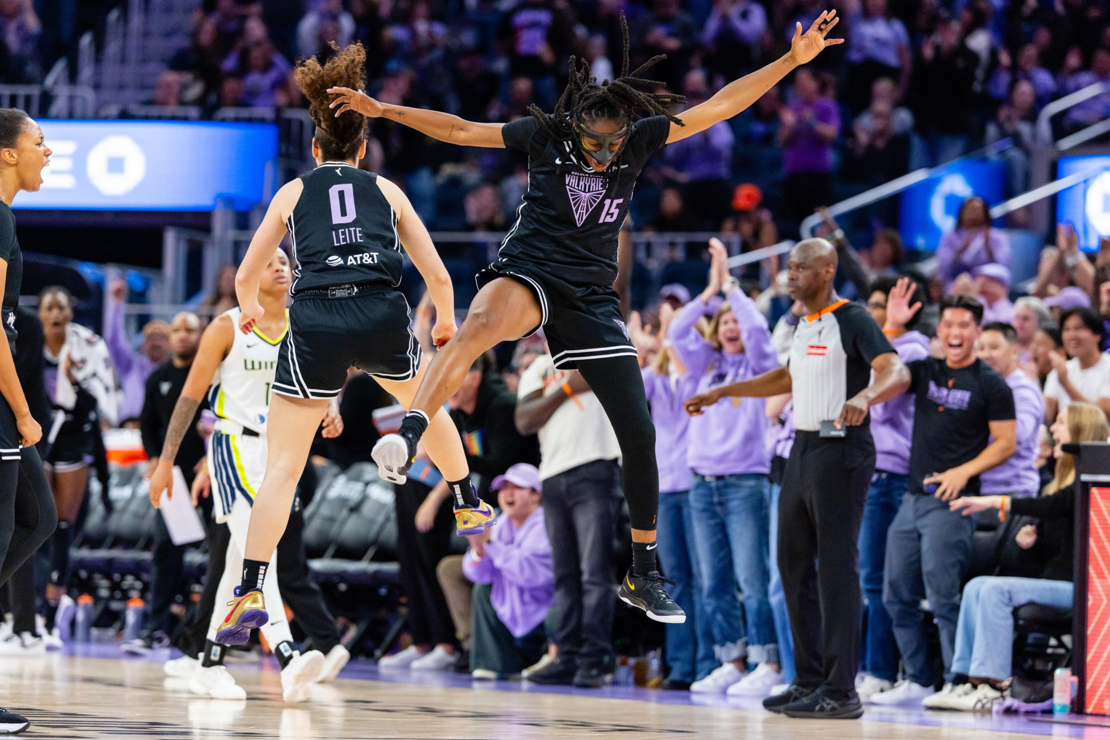 A basketball player in black jumps high, celebrating with teammates. The crowd in the background is cheering, creating an energetic atmosphere.