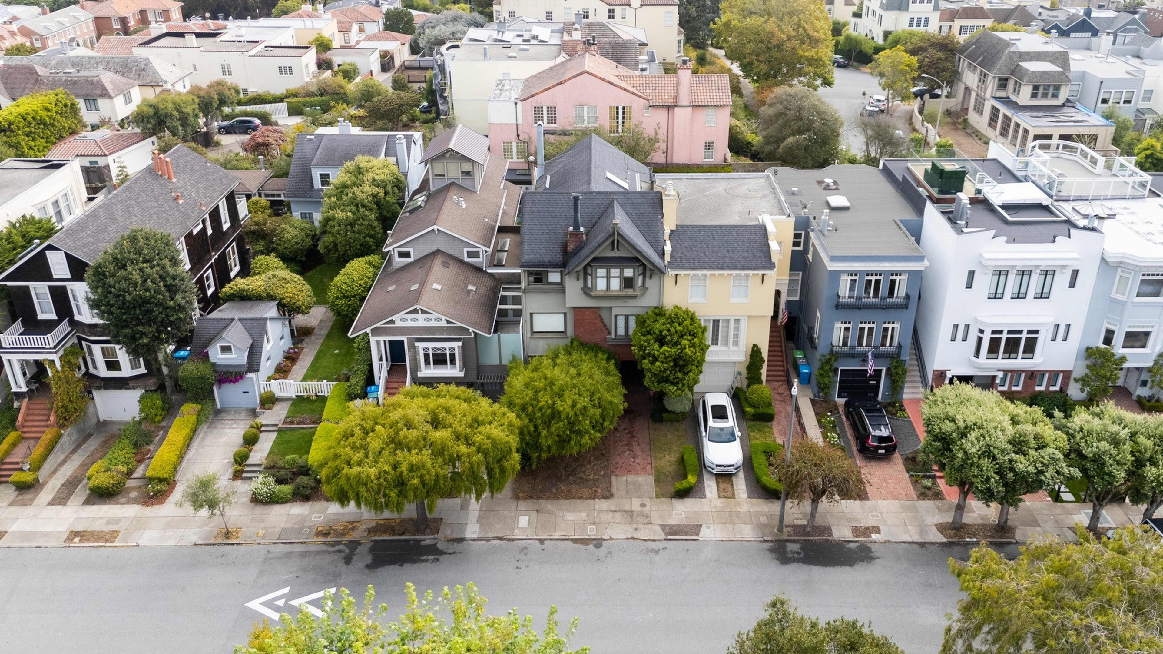 A row of closely spaced houses with varied architectural styles lines a tree-lined street, each with small front yards and driveways.