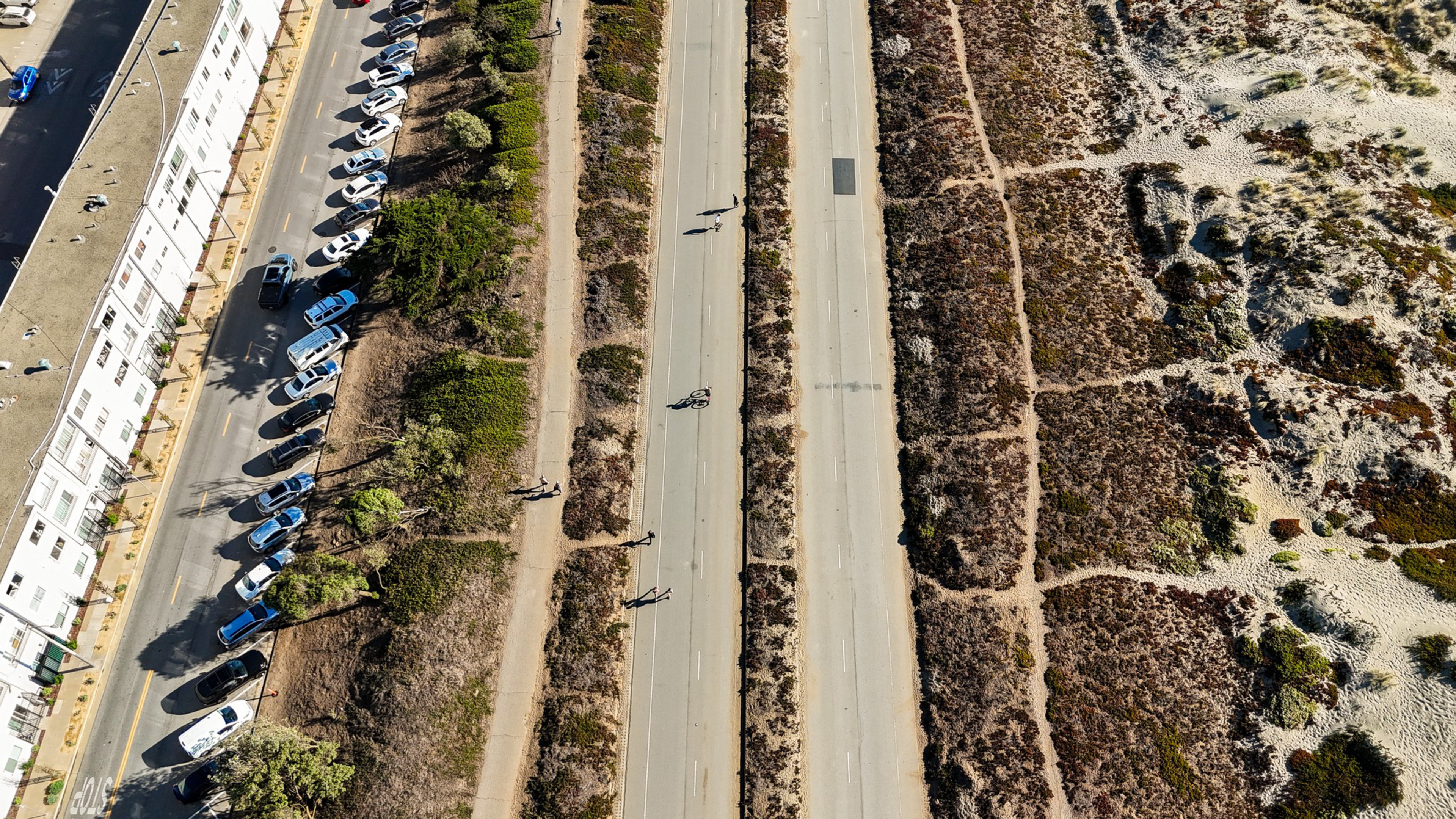 A wide paved road runs vertically with several people walking or cycling; parked cars line a building on the left, and grassy, sandy terrain lies on the right.