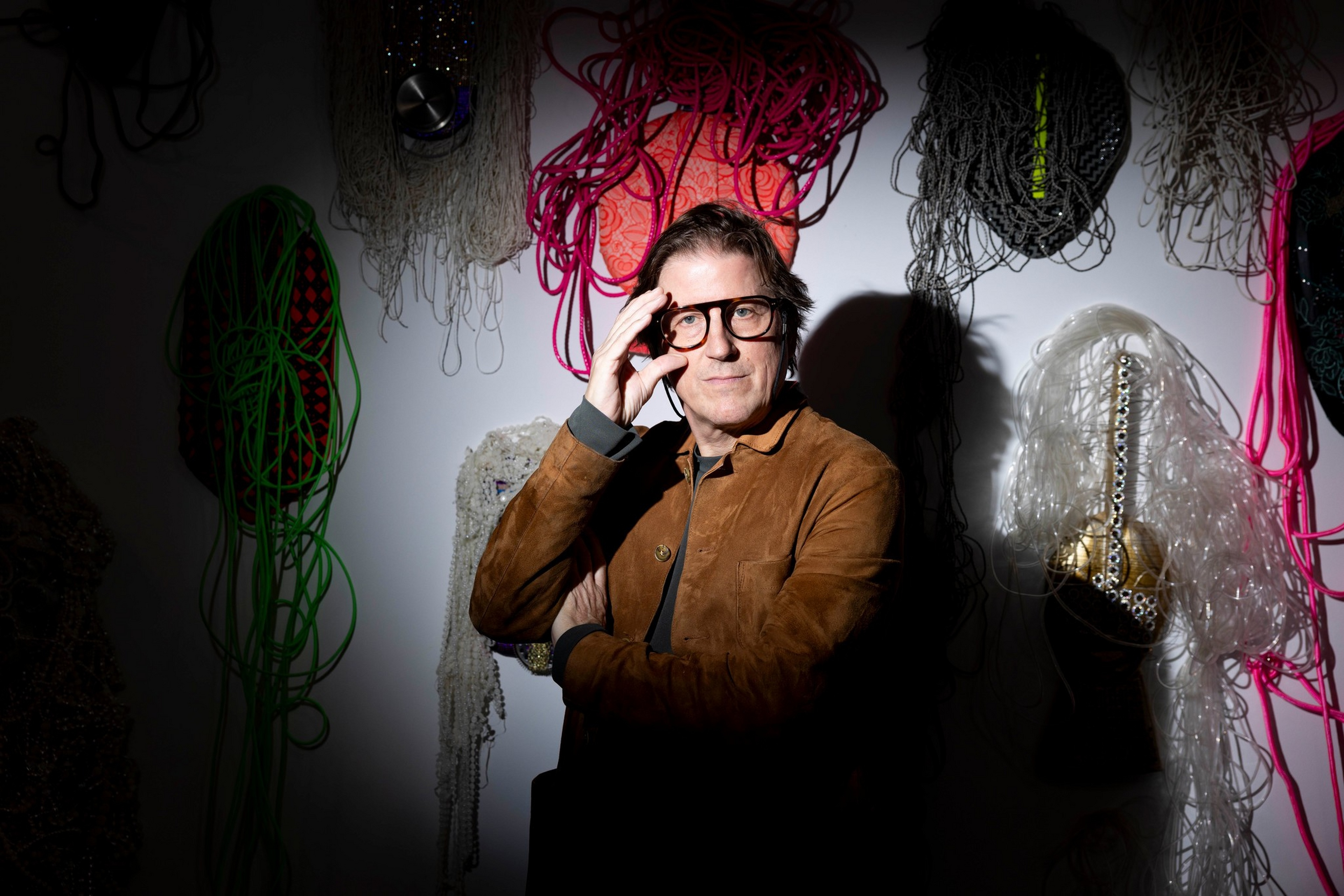 A man wearing glasses and a brown jacket poses thoughtfully against a wall decorated with colorful, textured hanging art pieces.