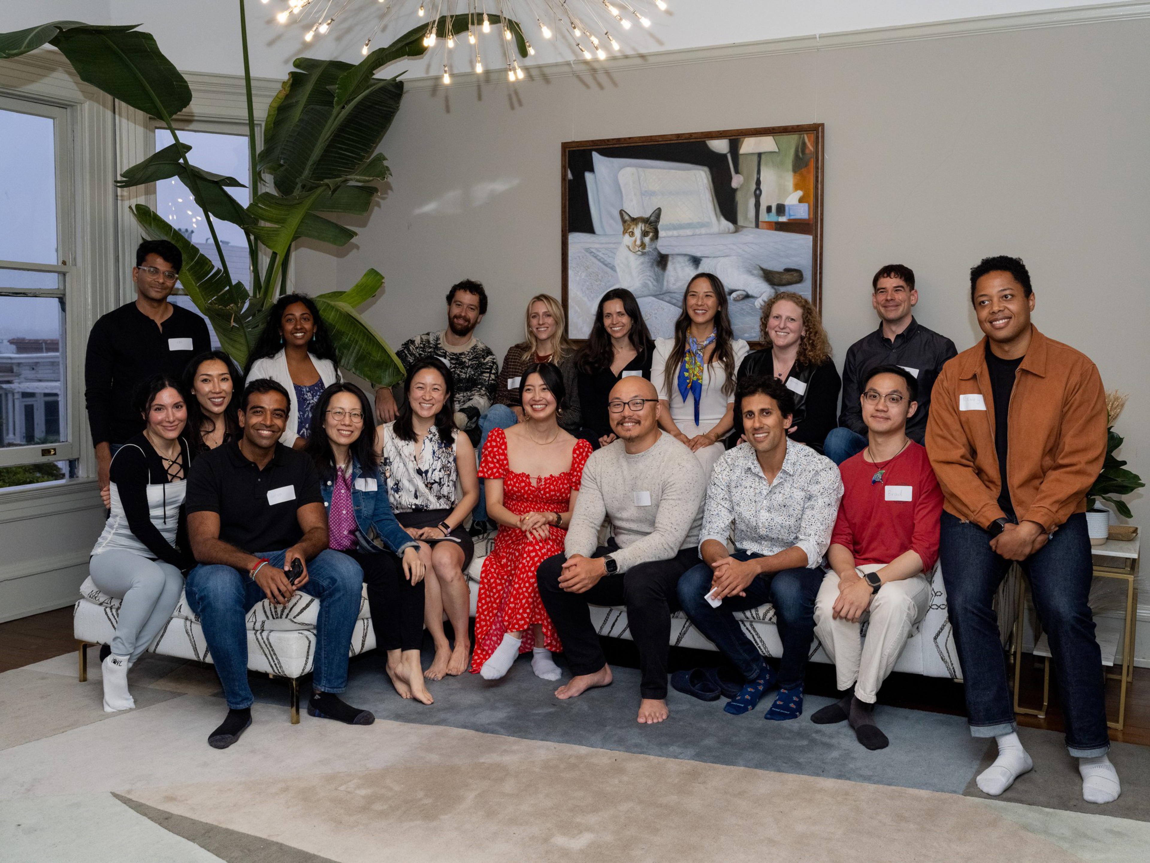 Fifteen diverse people smile, posing barefoot or socked in a cozy living room with large plants and a framed cat painting behind them.
