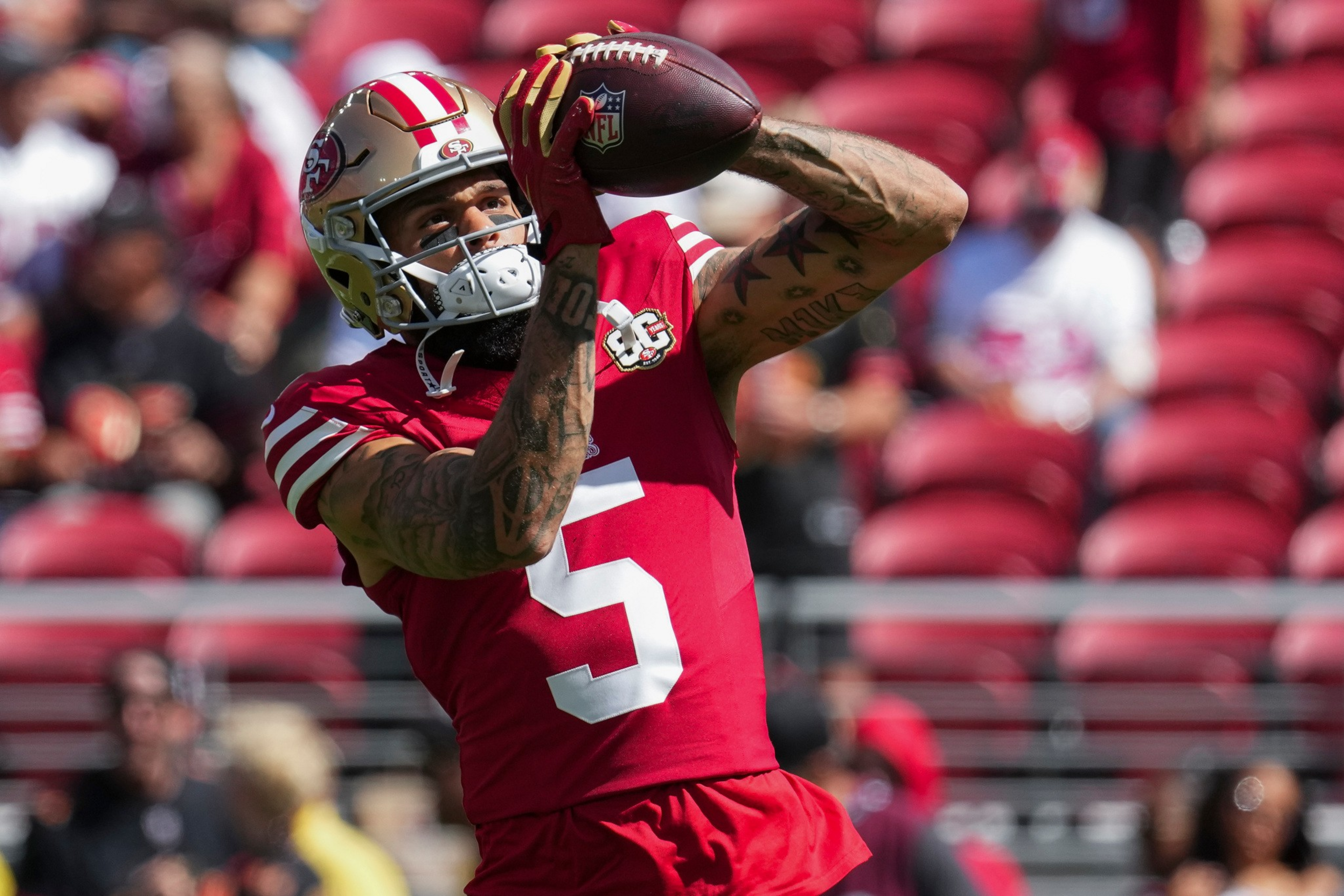 A football player in a red San Francisco 49ers uniform and gold helmet catches a football mid-air in a stadium with blurred spectators.