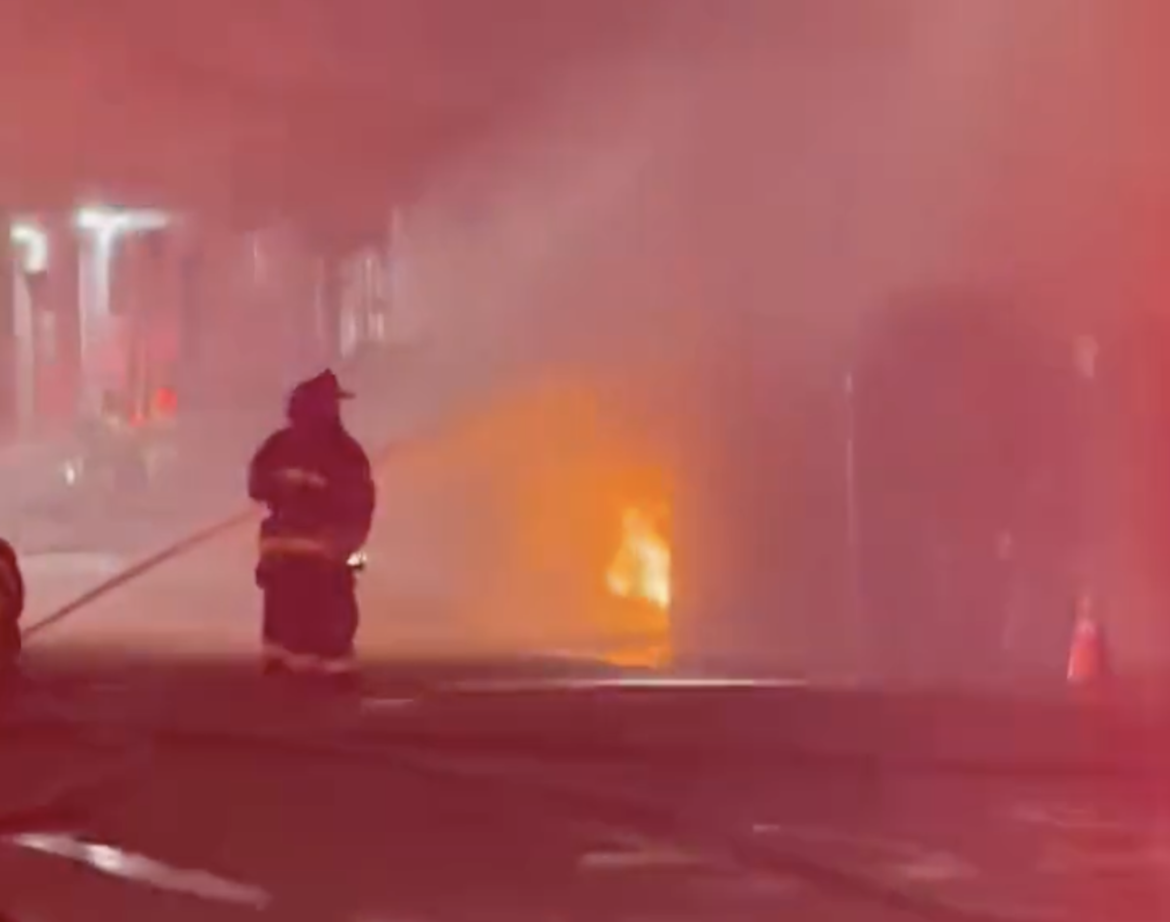 A firefighter is spraying water on a fire engulfing a structure, with red emergency lights illuminating the smoky scene.