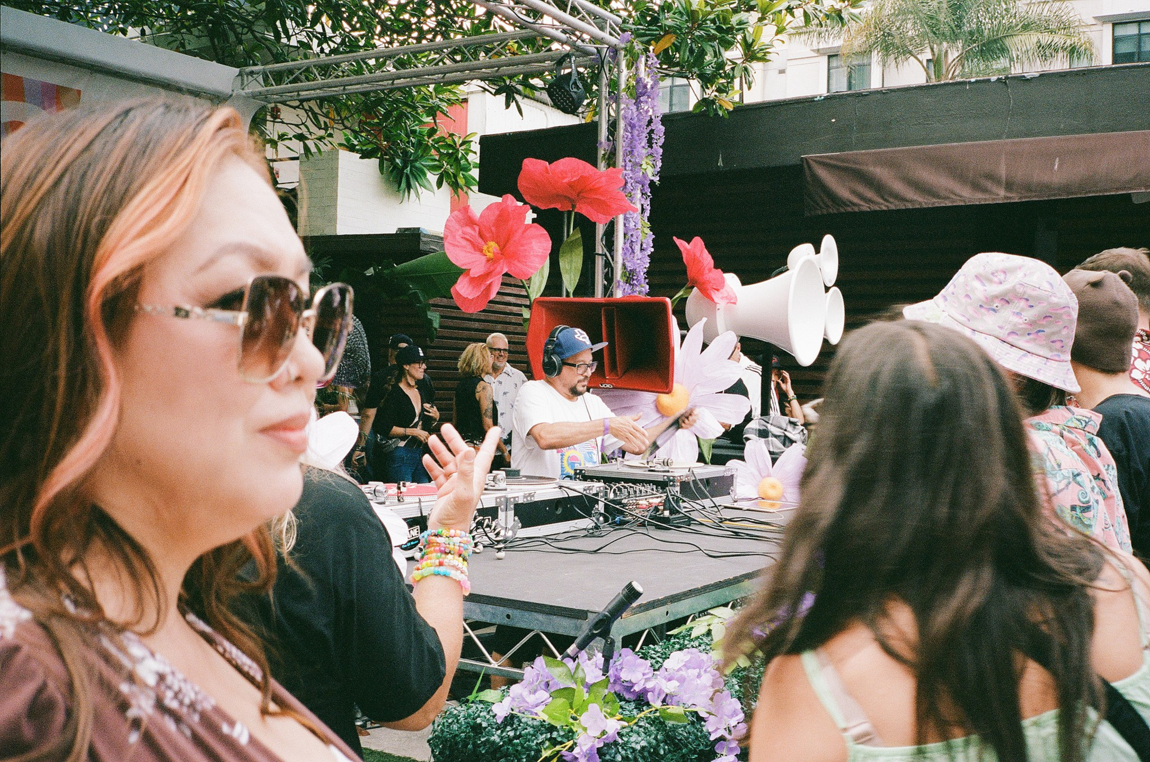 A DJ wearing headphones and a cap plays music at a decorated outdoor event with colorful flowers and people enjoying the scene.