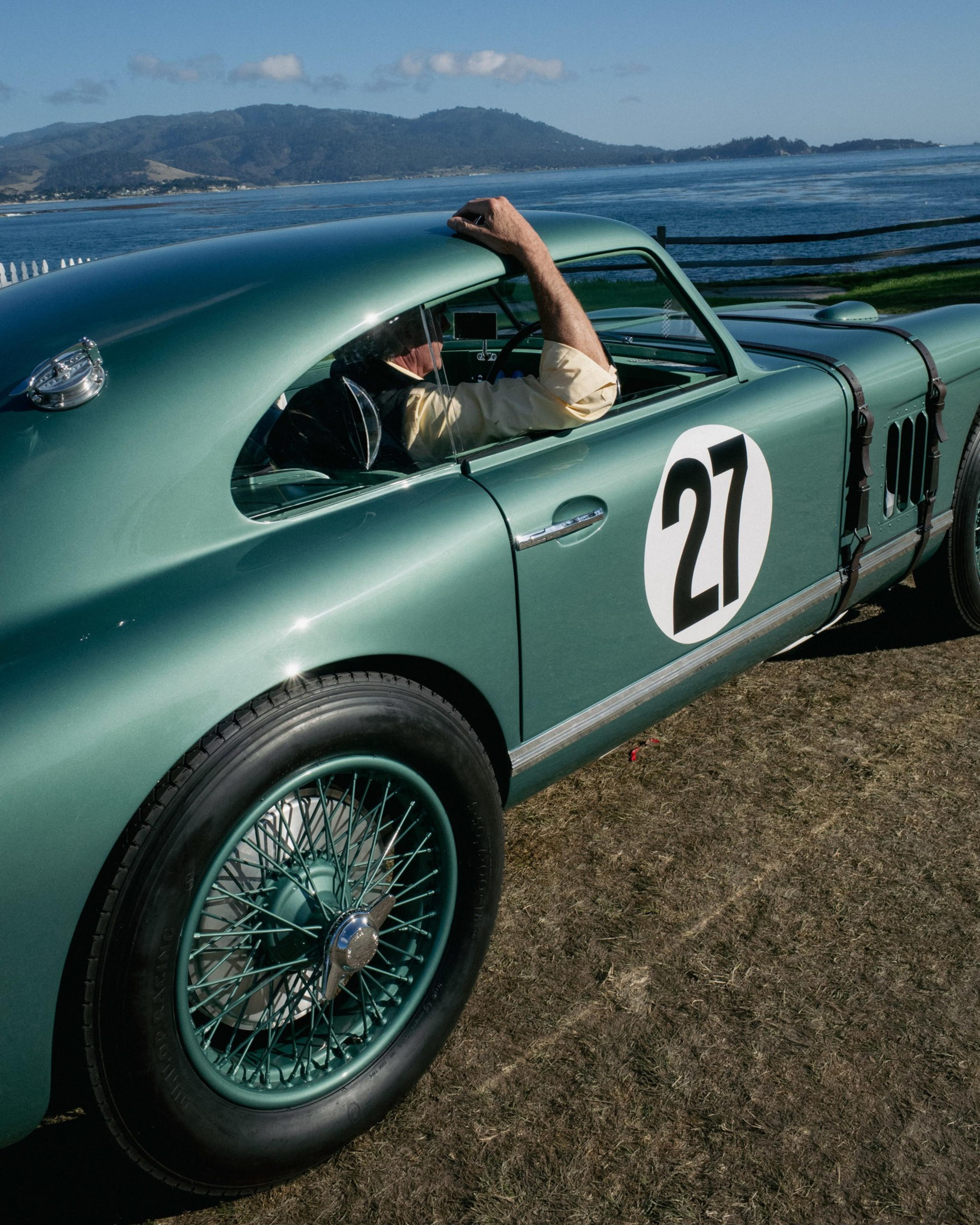 A vintage green race car with the number 27 on its side is parked by a waterfront, with a driver resting their arm on the window frame.