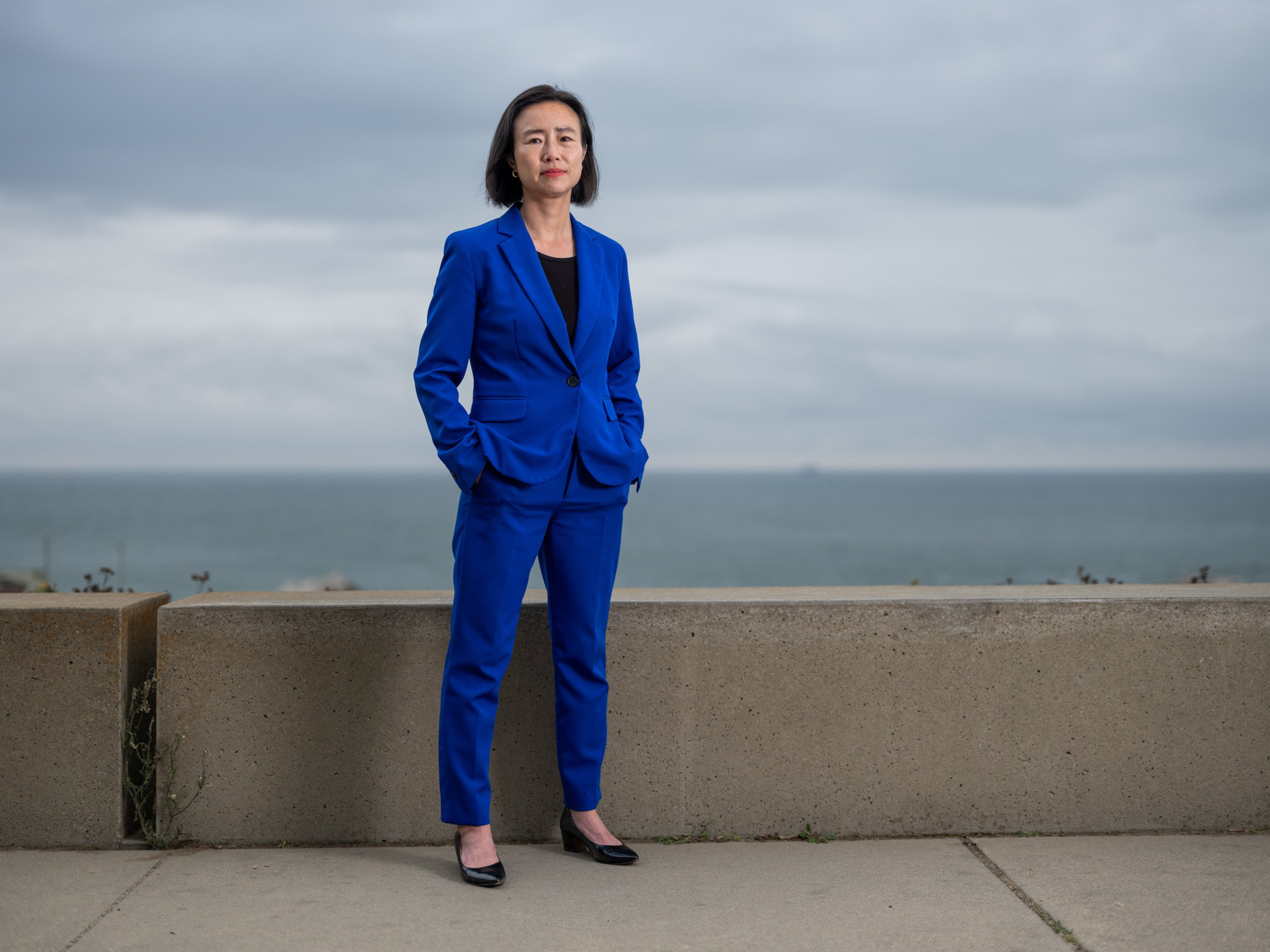 A woman in a blue suit and black shoes stands confidently with hands in pockets by a concrete barrier, with the ocean and cloudy sky in the background.