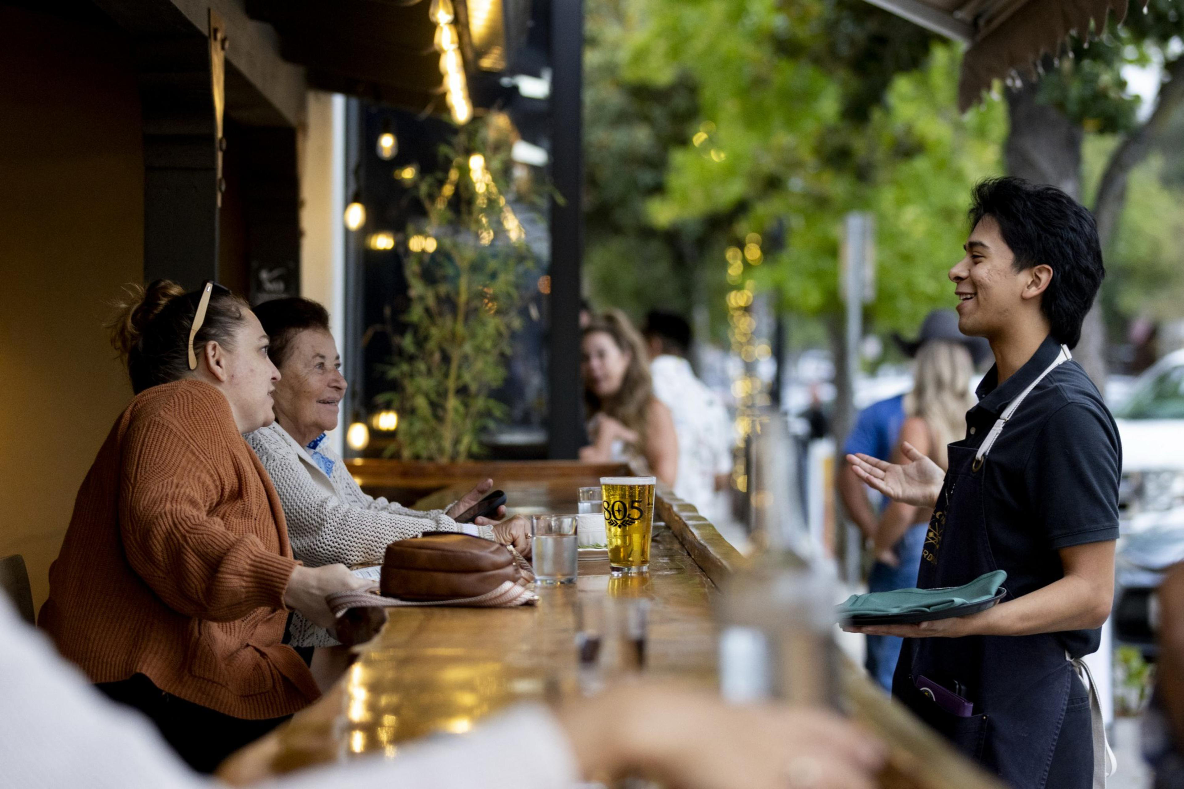 Two women sit at a bar talking to a smiling waiter holding a tray with outdoor trees and string lights in the background.