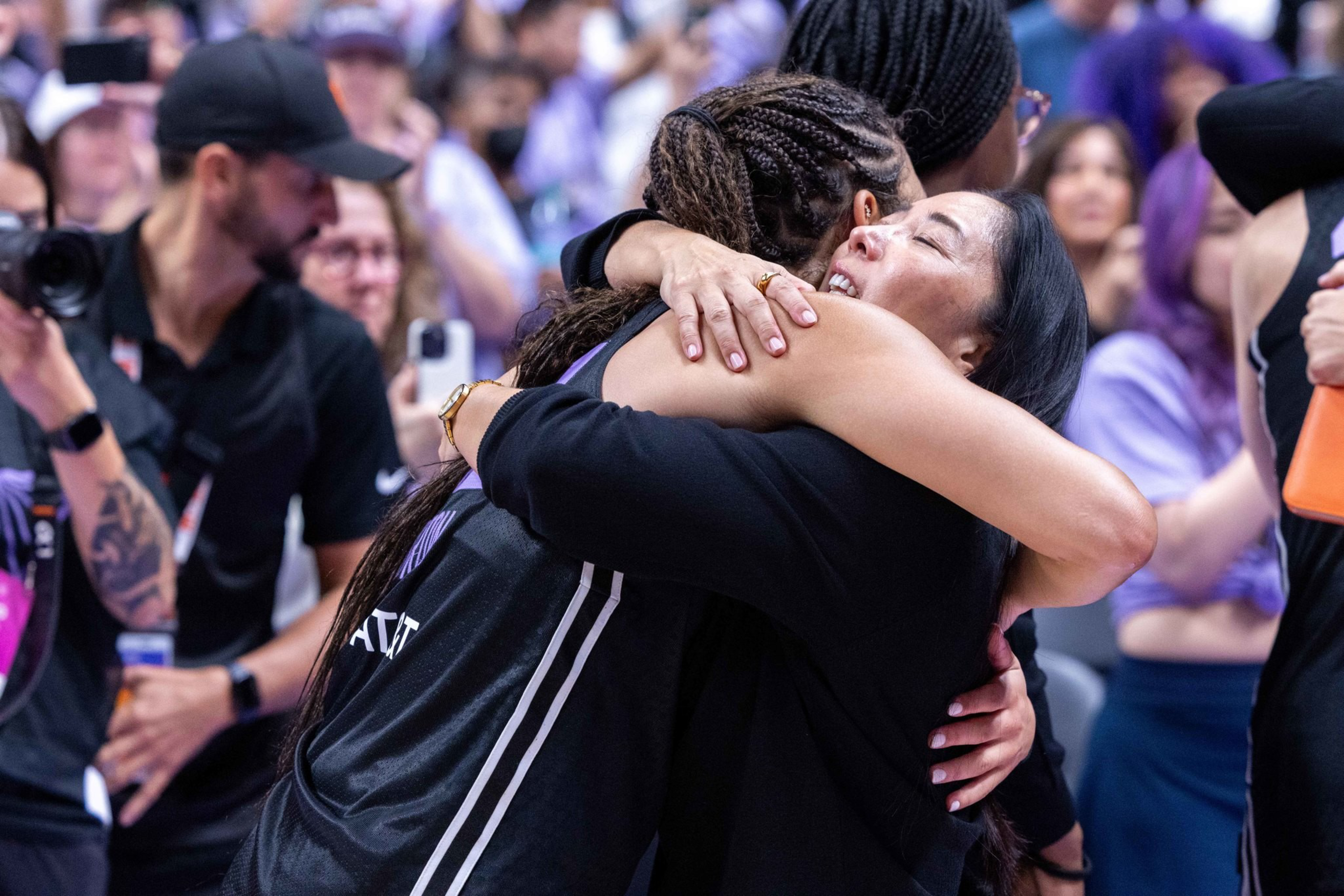 A basketball player in a black jersey embraces a woman warmly, with a joyful crowd blurred in the background.