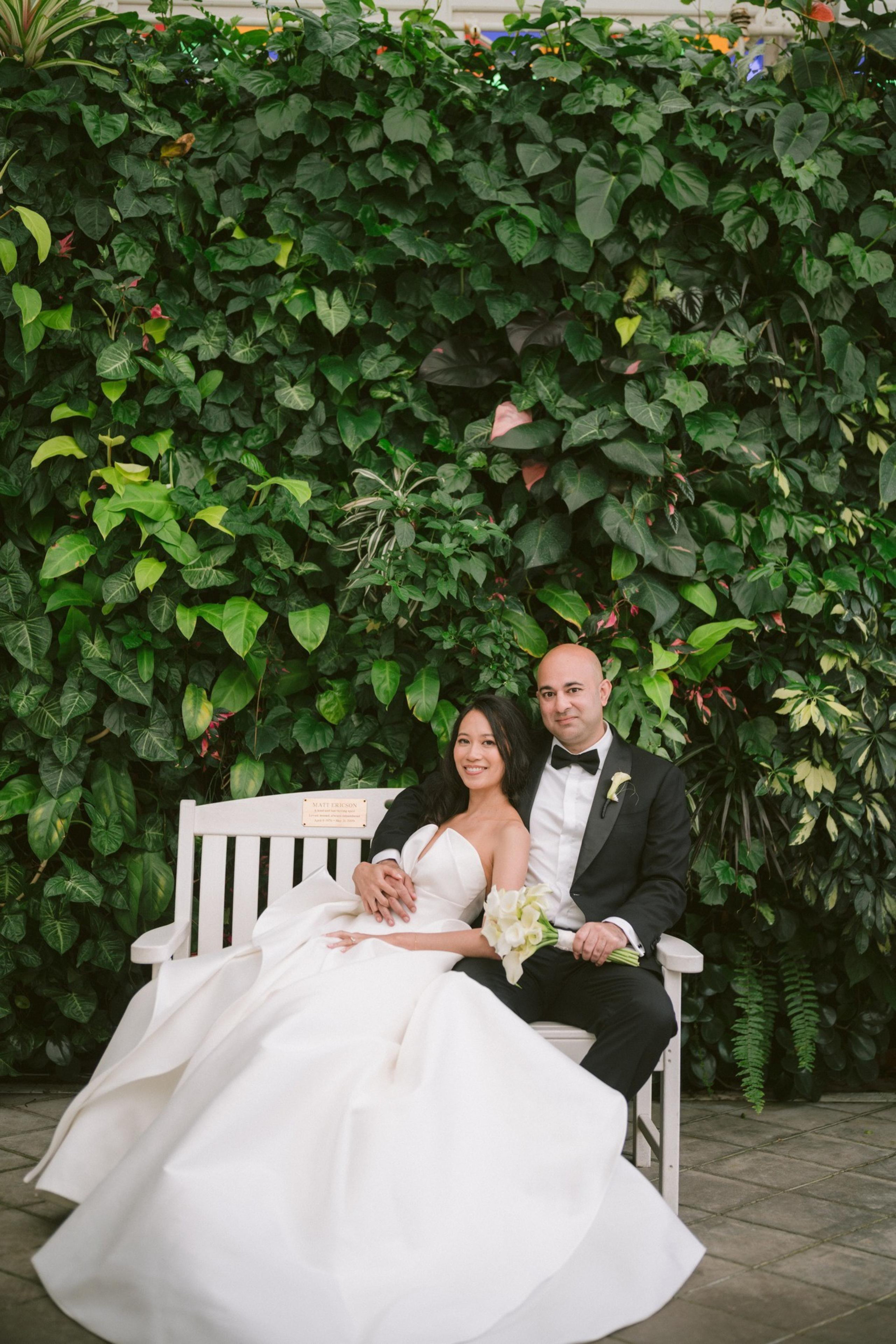 A bride in a white gown and a groom in a black tuxedo sit closely on a white bench, surrounded by lush green foliage.