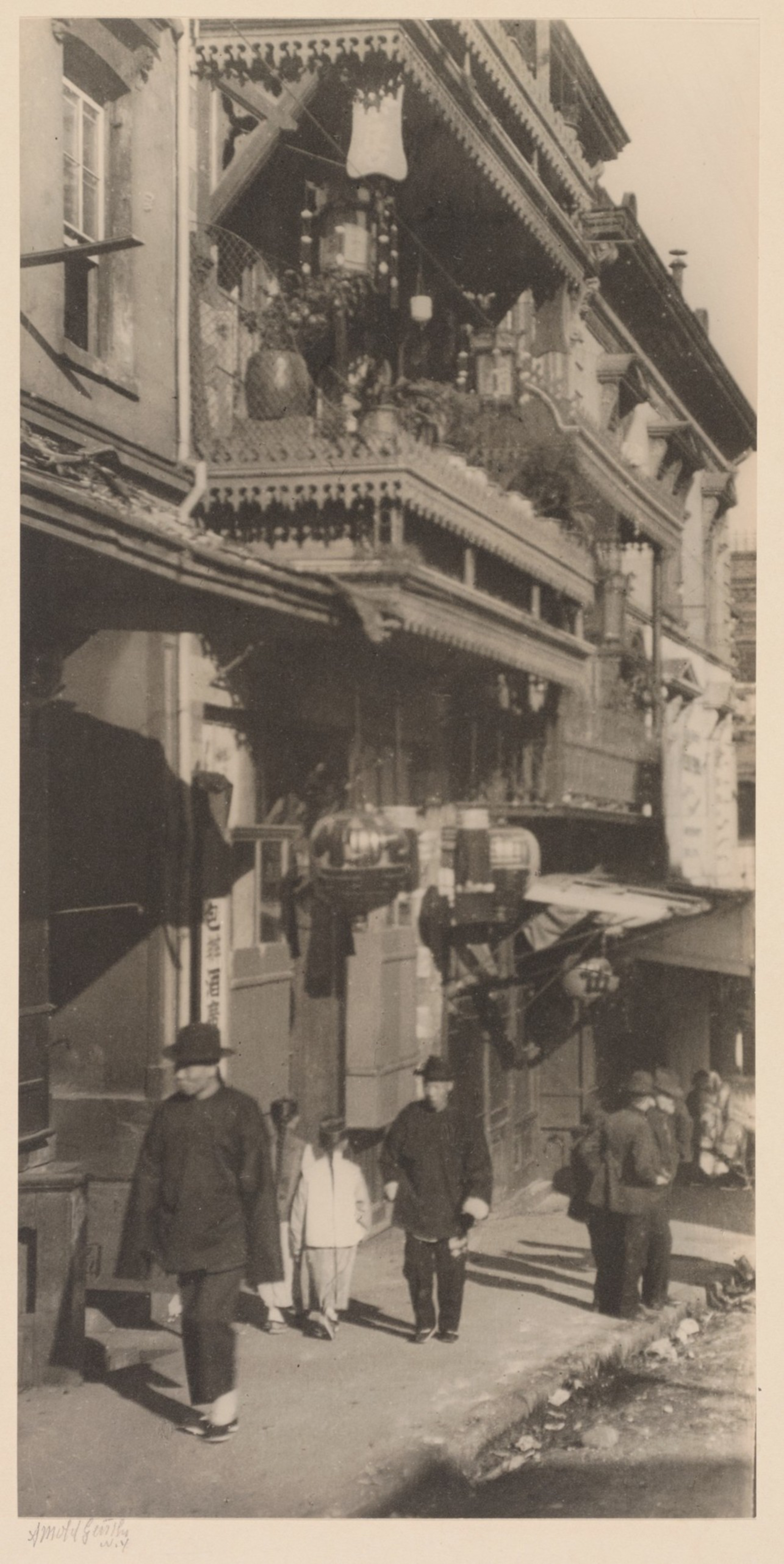 A narrow street with traditional Asian-style buildings, decorated balconies, and people walking, some dressed in early 20th-century attire.