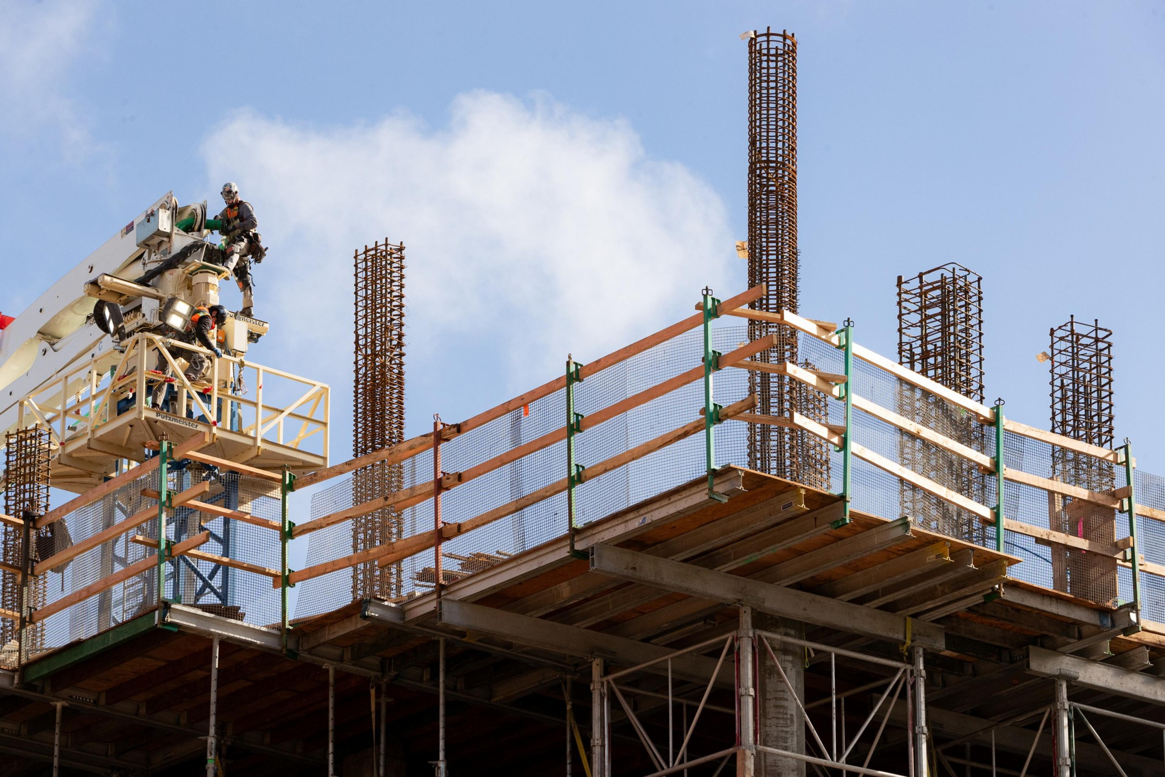 Two construction workers on a lift work on a building's upper floor with exposed steel rebar columns and temporary safety railings.