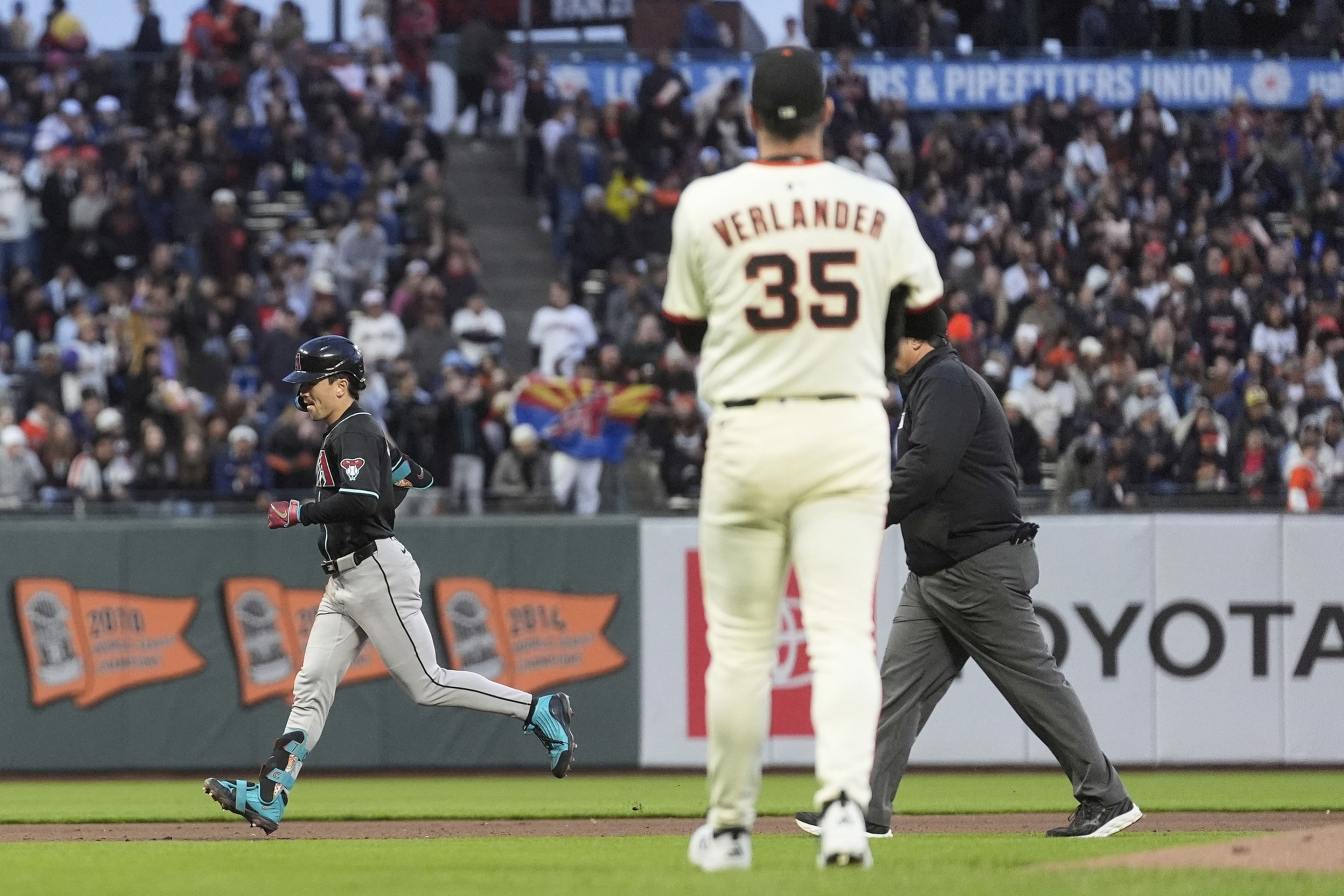 A baseball player in a black jersey runs past a pitcher in a white jersey with &quot;35&quot; on the back. An umpire and a crowd with banners are in the background.