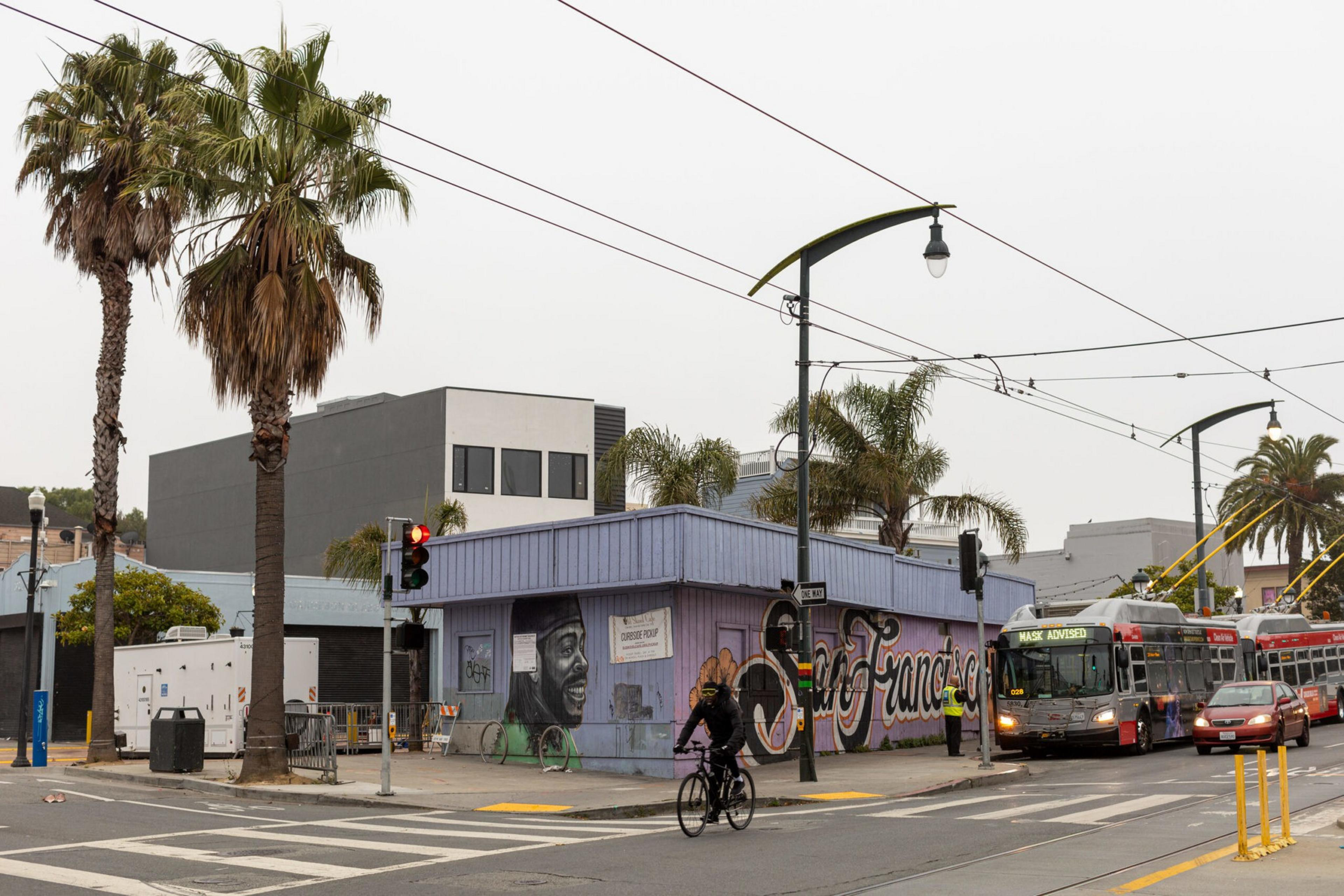A cyclist rides past a corner building with a mural and palm trees, while a San Francisco bus waits at a traffic light under an overcast sky.