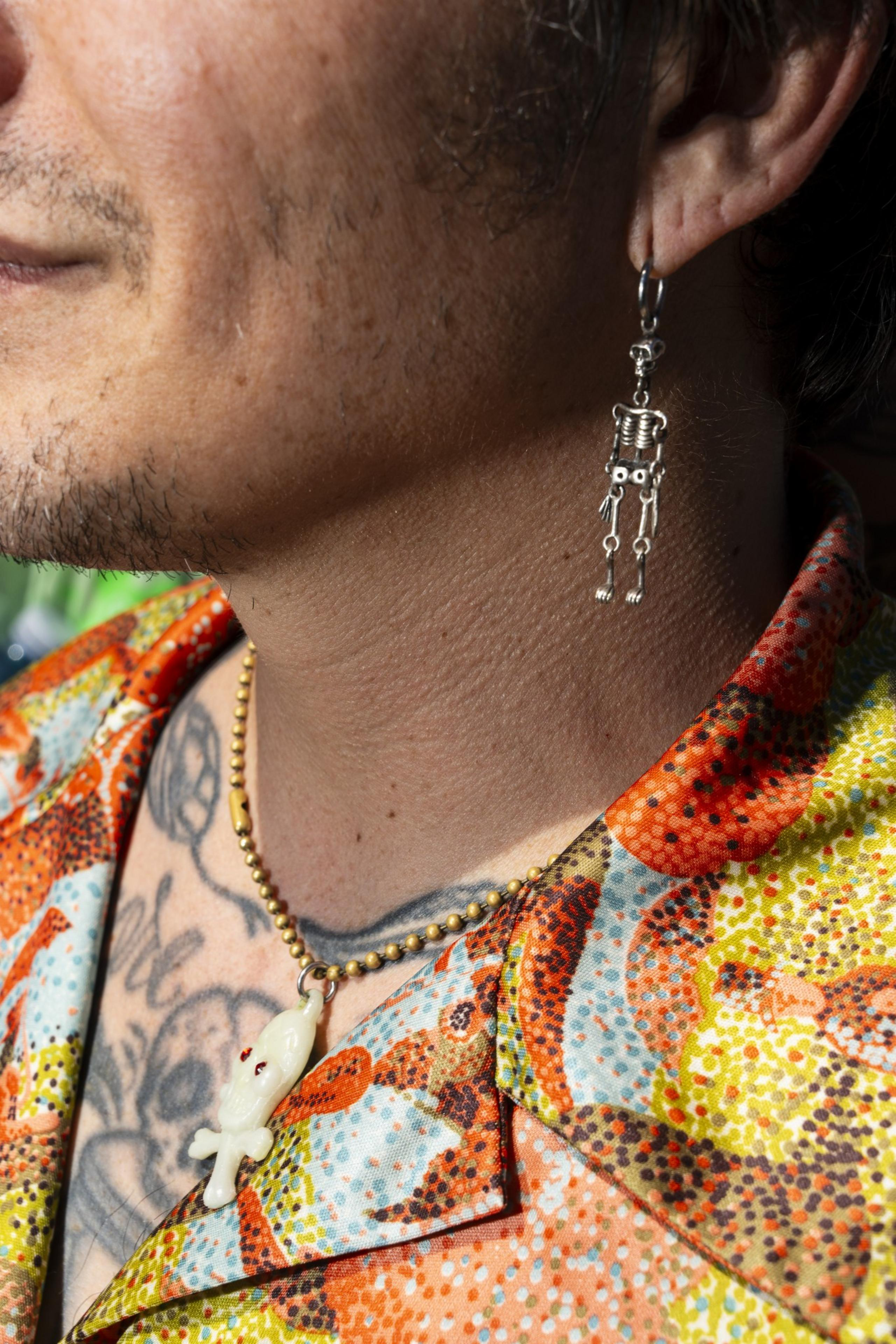 A person wears a colorful, patterned shirt with tattoos visible on their chest. They have a skeleton earring and a necklace with a small alien pendant.