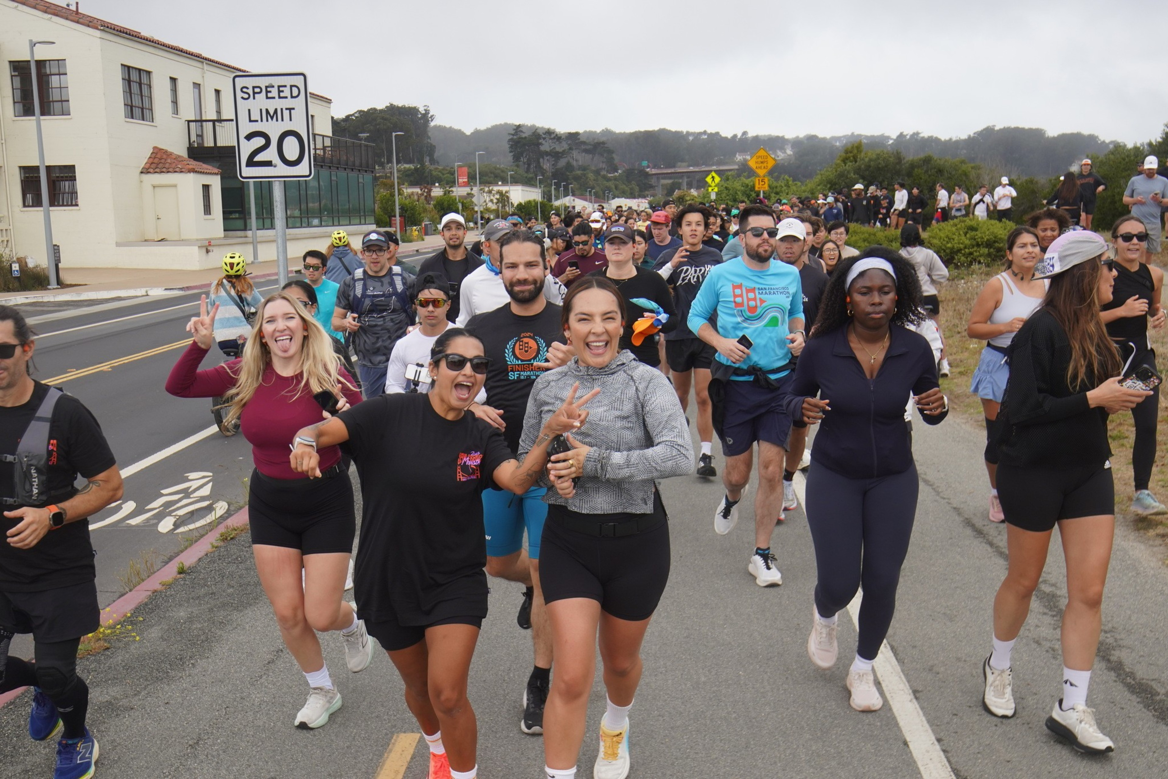 A group of diverse runners smiling energetically as they run together on a road, surrounded by scenic greenery and buildings, near a 20 mph speed limit sign.