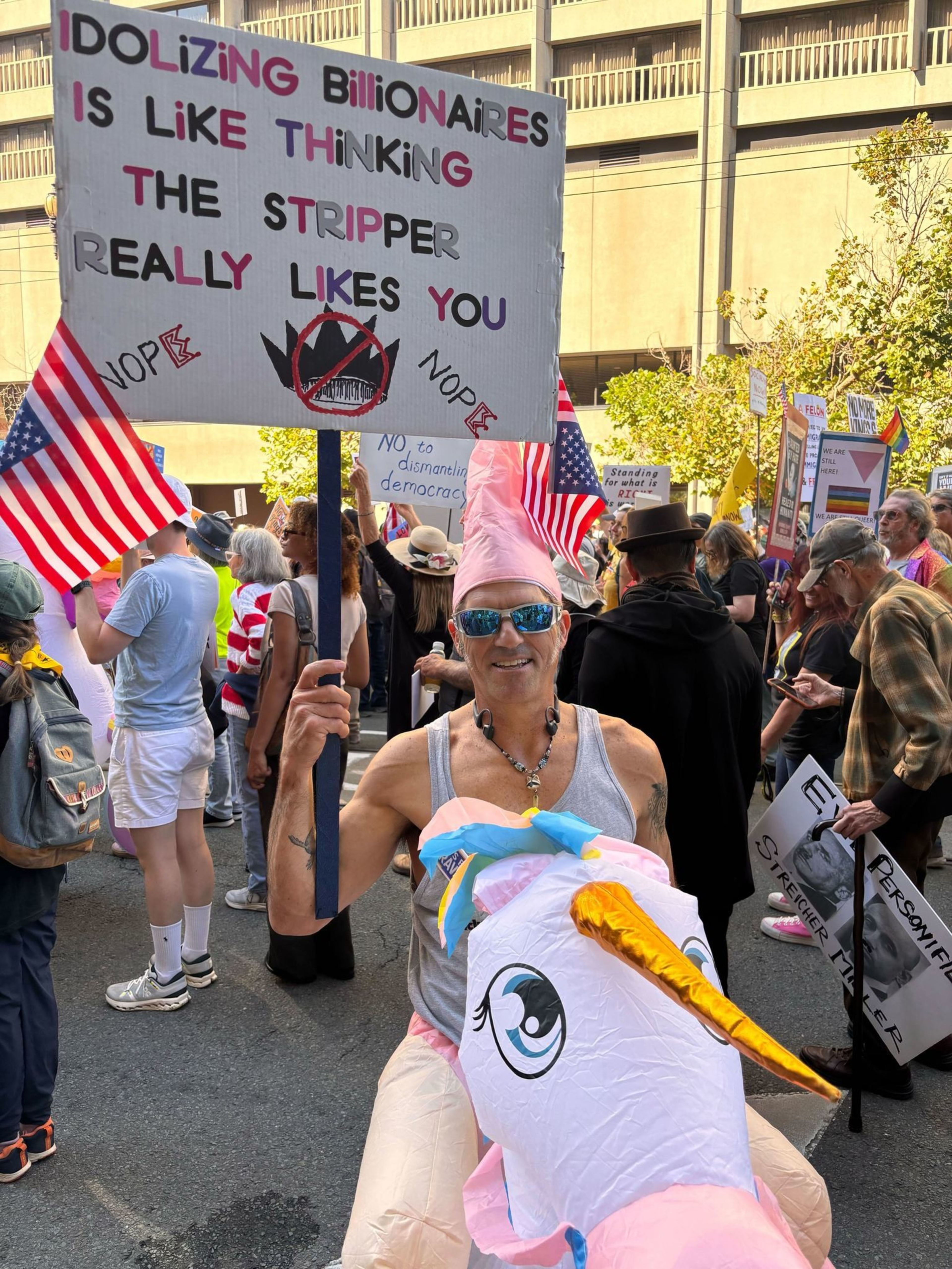 A man in sunglasses and a pink hat rides an inflatable unicorn while holding a protest sign against idolizing billionaires. Several people hold American flags nearby.