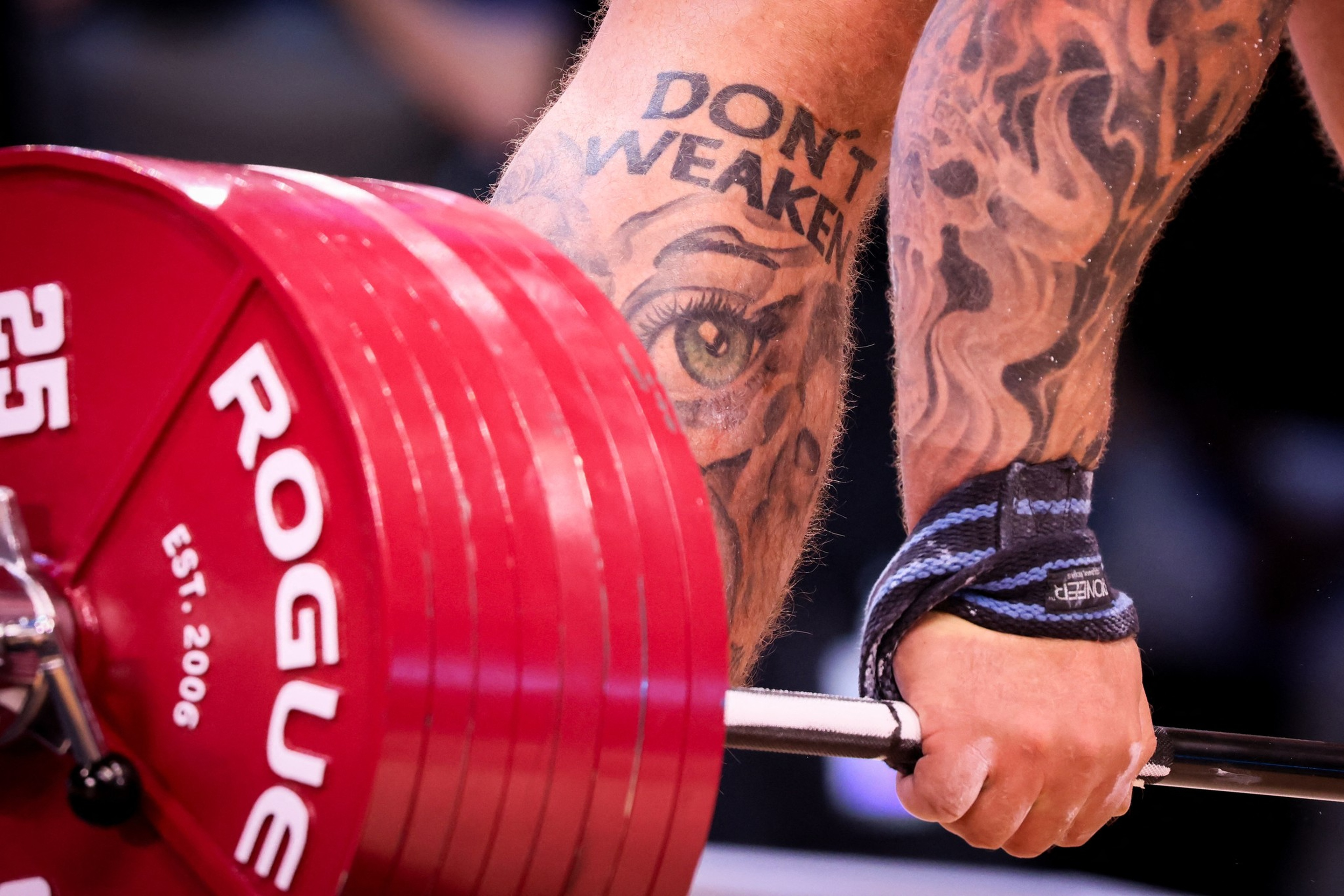 A heavily tattooed arm with “DON’T WEAKEN” text lifts a barbell loaded with large red Rogue weight plates. The wrist has a blue and black lifting strap.