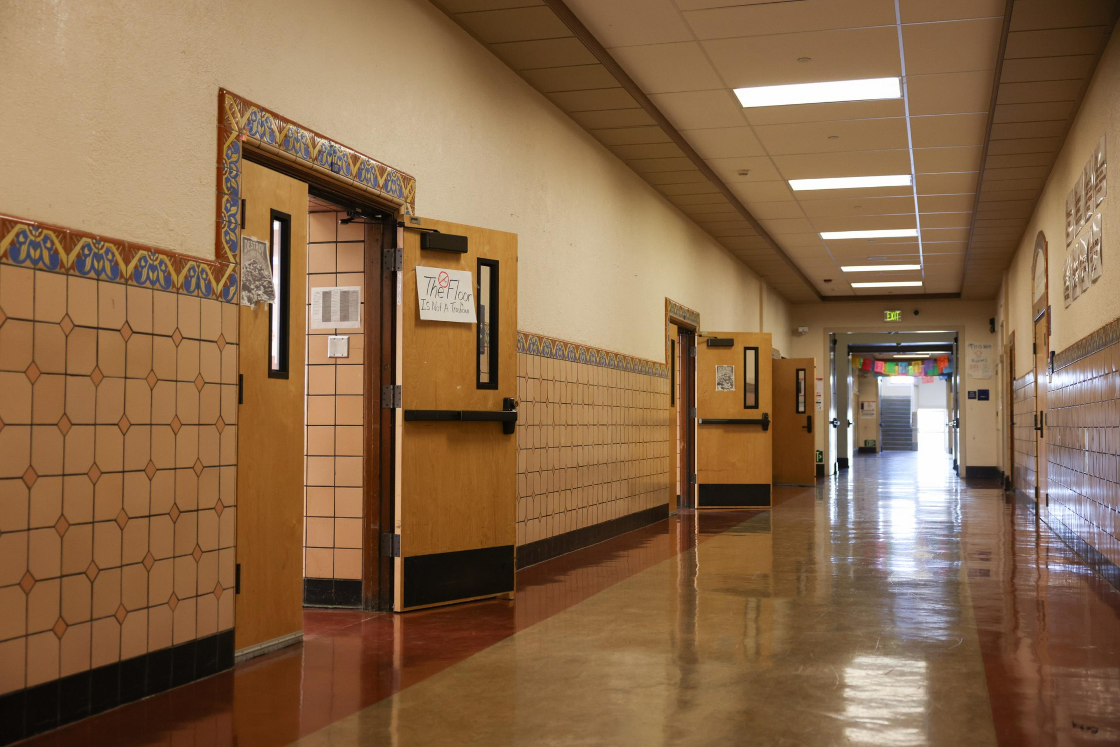 A shiny school hallway with beige tiled walls, open wooden classroom doors, fluorescent ceiling lights, and colorful flags visible at the far end.