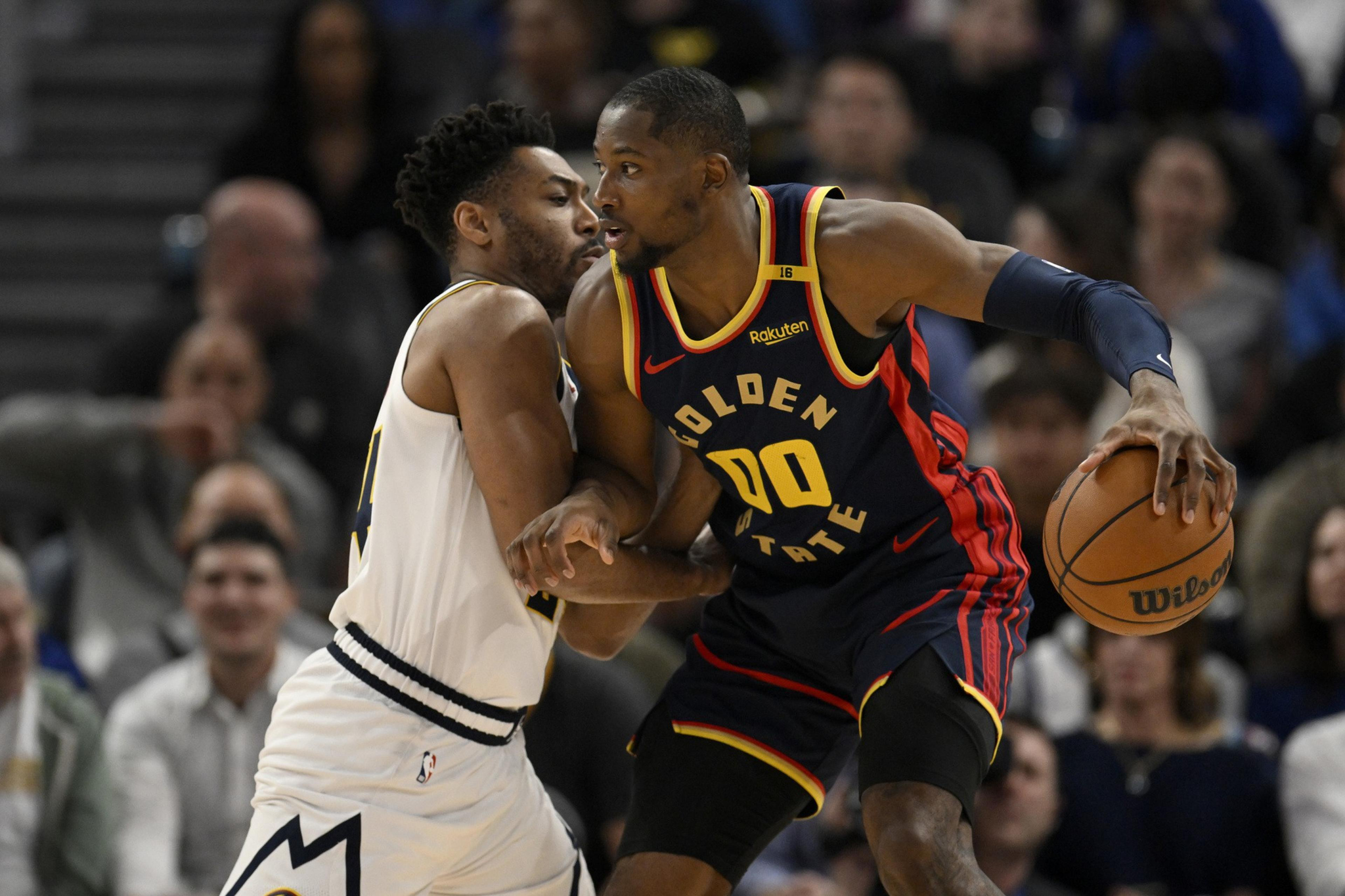 A Golden State Warriors player in navy and yellow holds a basketball while closely guarded by a defender in white during a game.