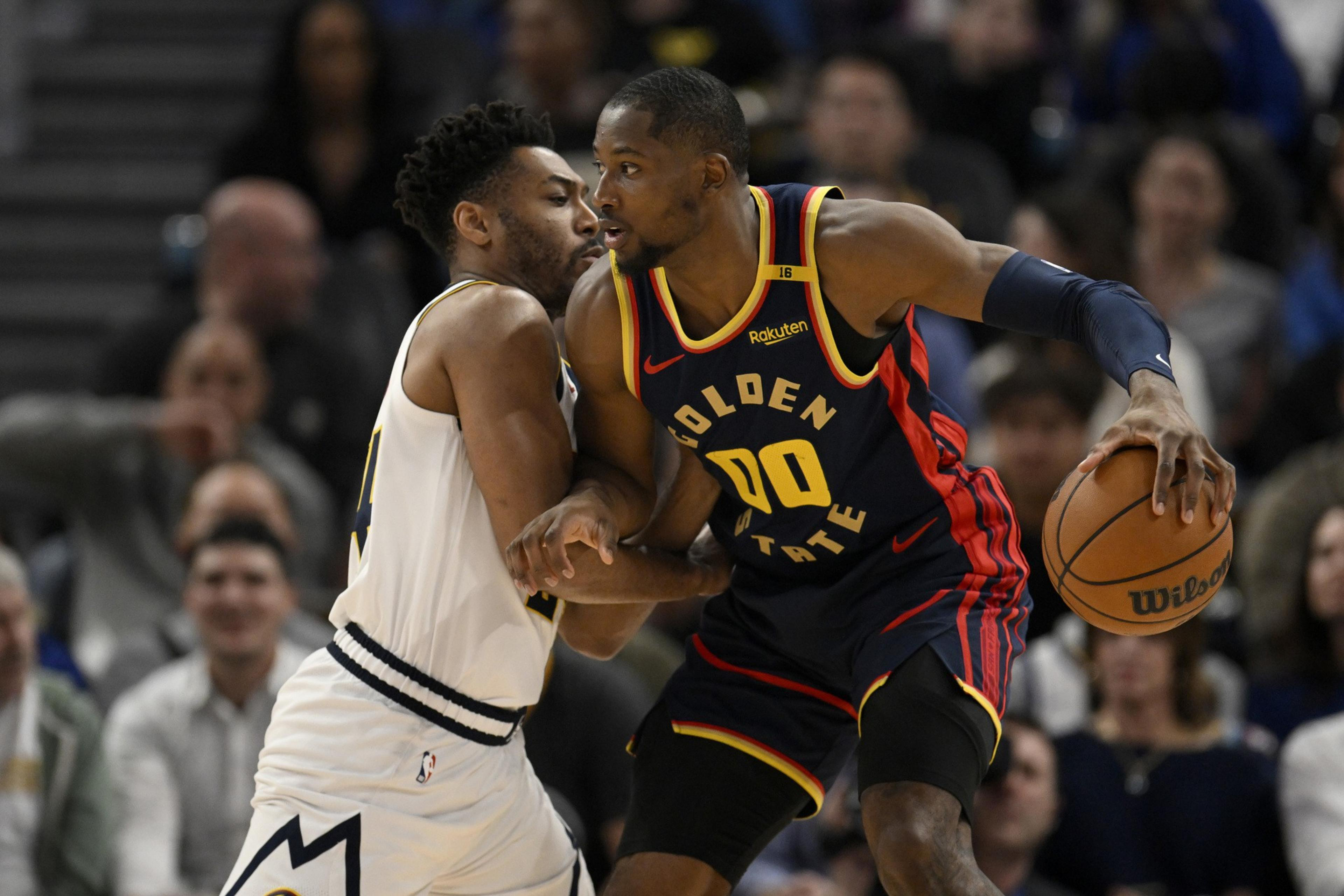 A Golden State Warriors player in navy and yellow holds a basketball while closely guarded by a defender in white during a game.