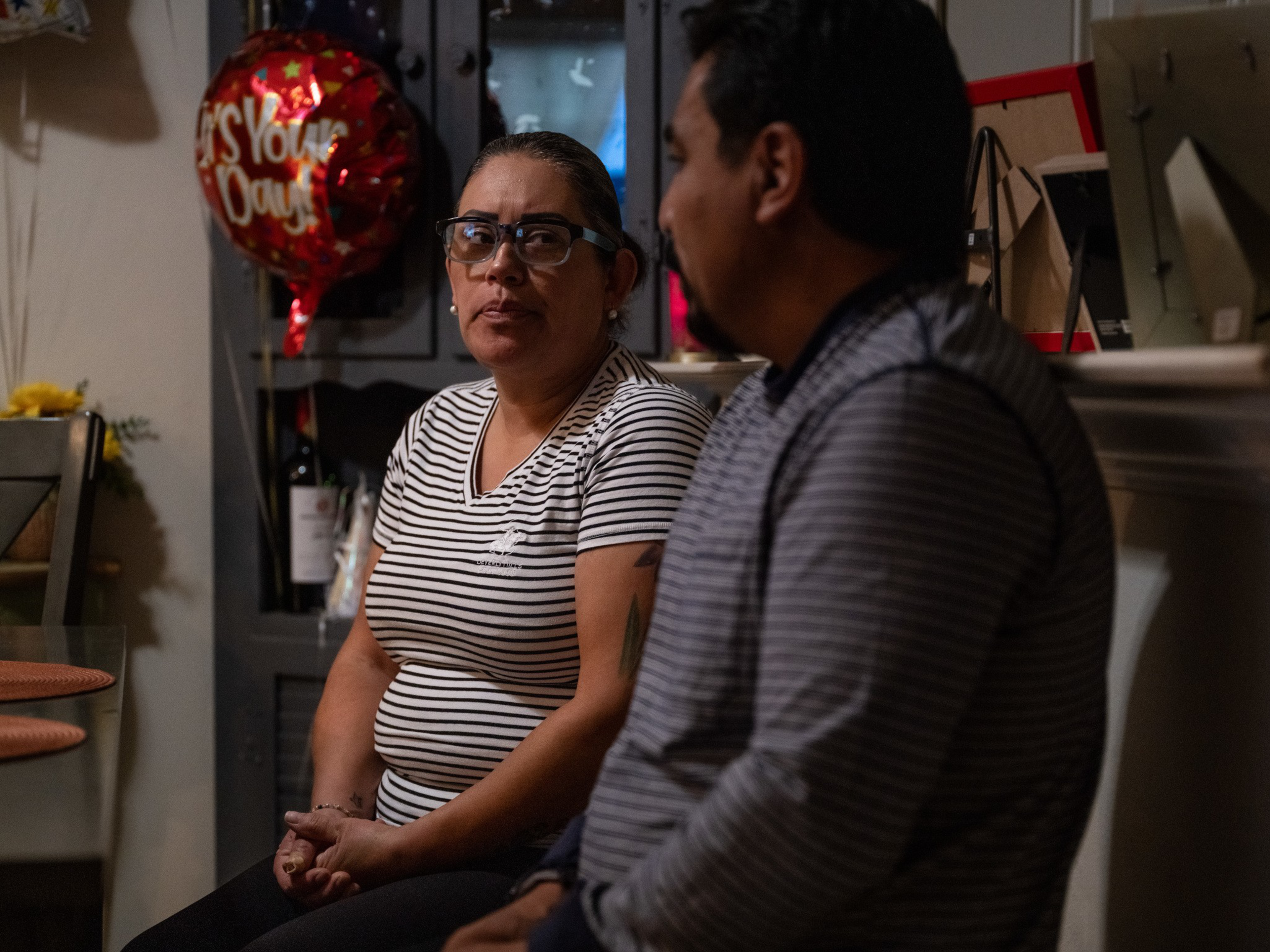 A woman wearing glasses and a striped shirt sits next to a man in a striped sweater, with a red balloon saying “It’s Your Day” behind them.