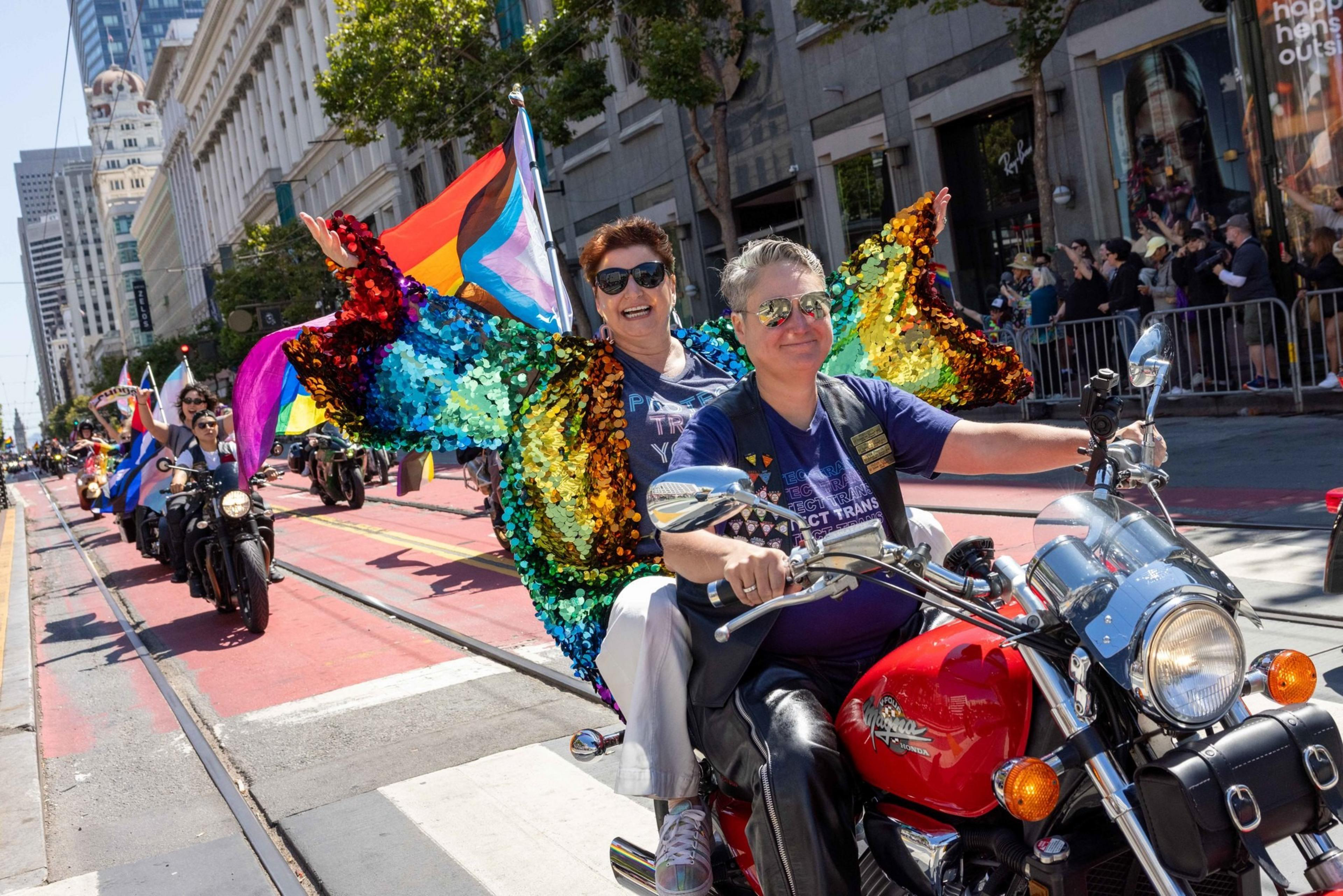 Two people ride a motorcycle in a parade, with one wearing a colorful sequin outfit and holding a Pride flag. Other motorcycles and spectators are visible.
