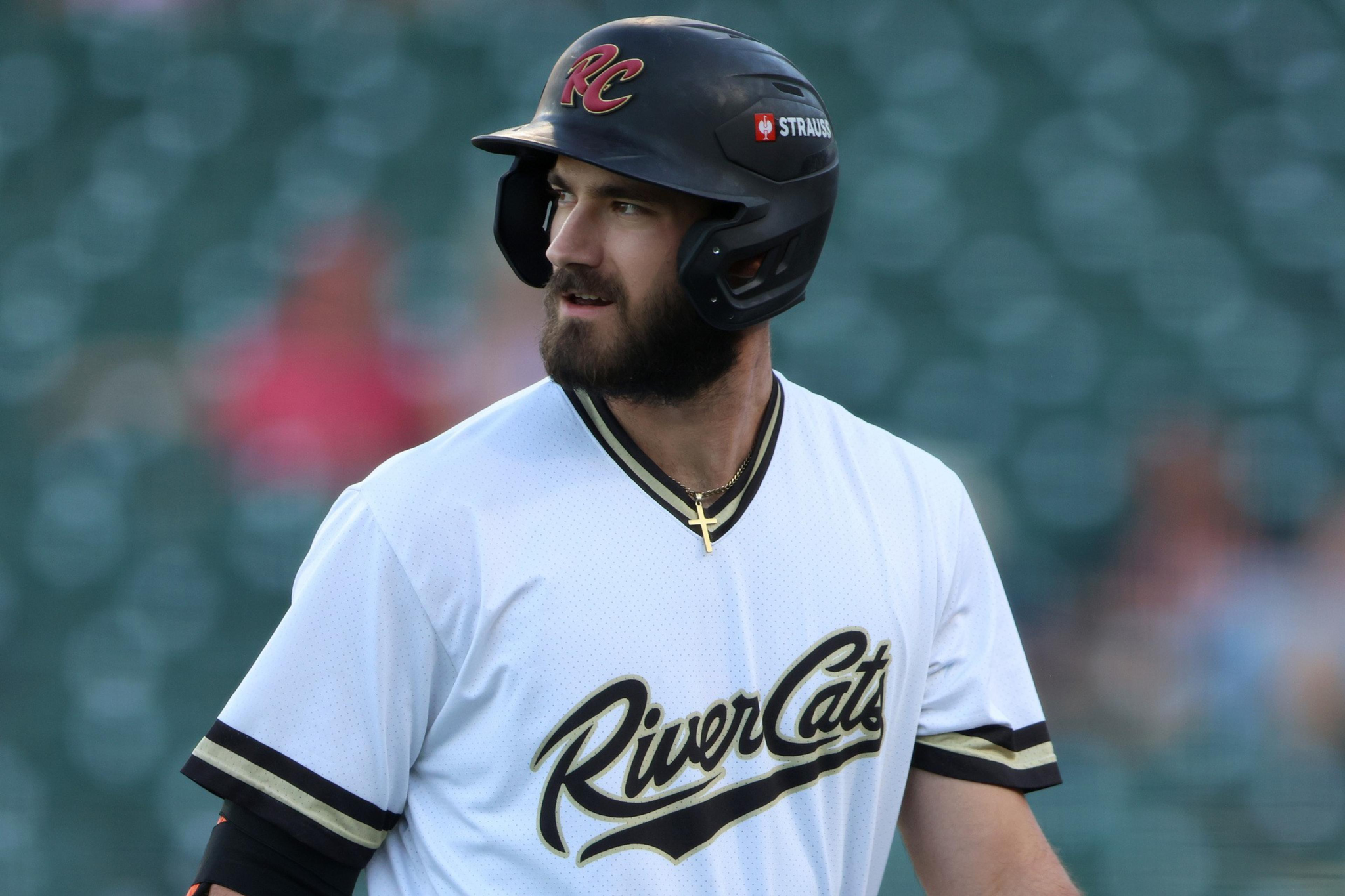 A bearded baseball player wearing a white jersey with "River Cats" written on it and a black helmet looks to his left.
