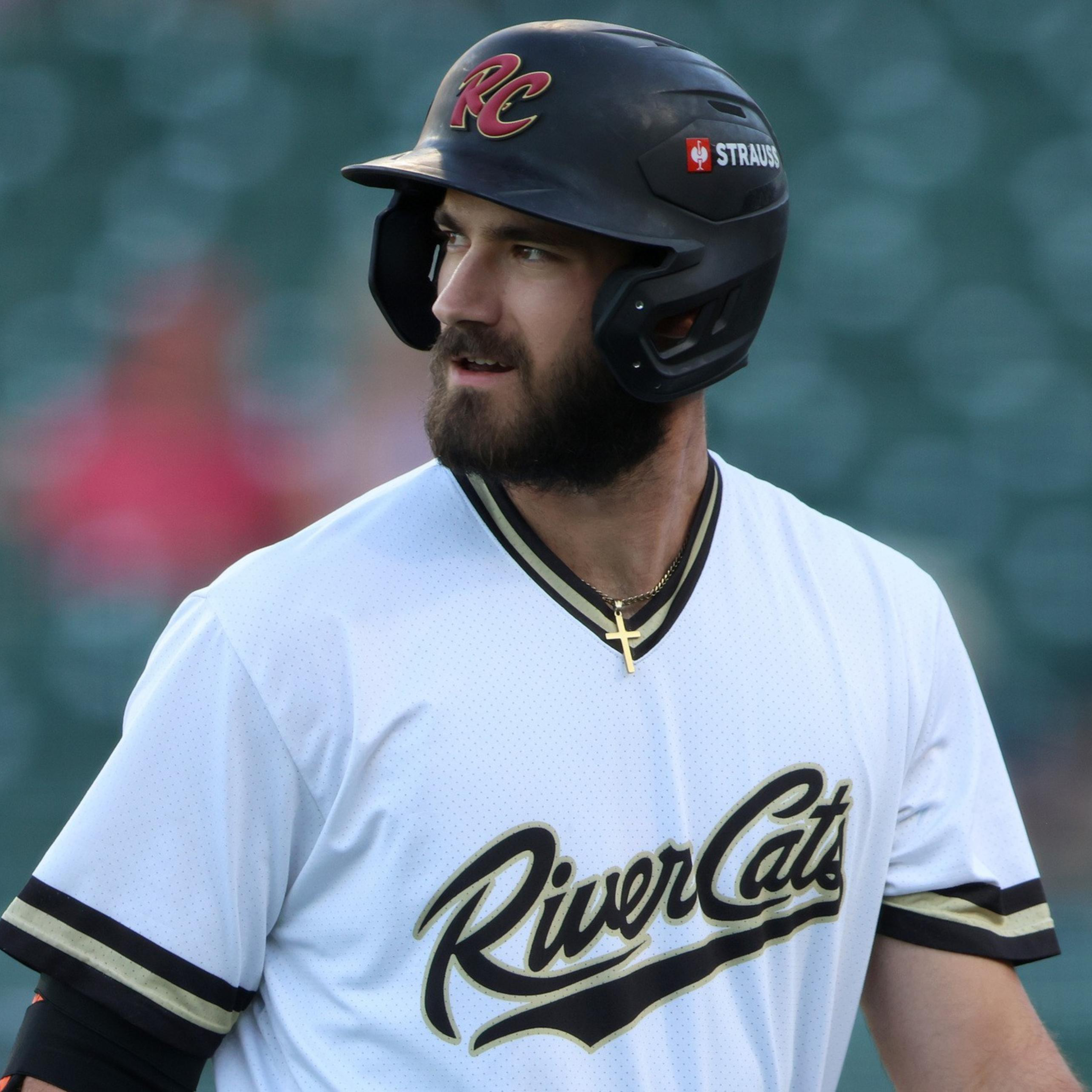 A bearded baseball player wearing a white jersey with "River Cats" written on it and a black helmet looks to his left.