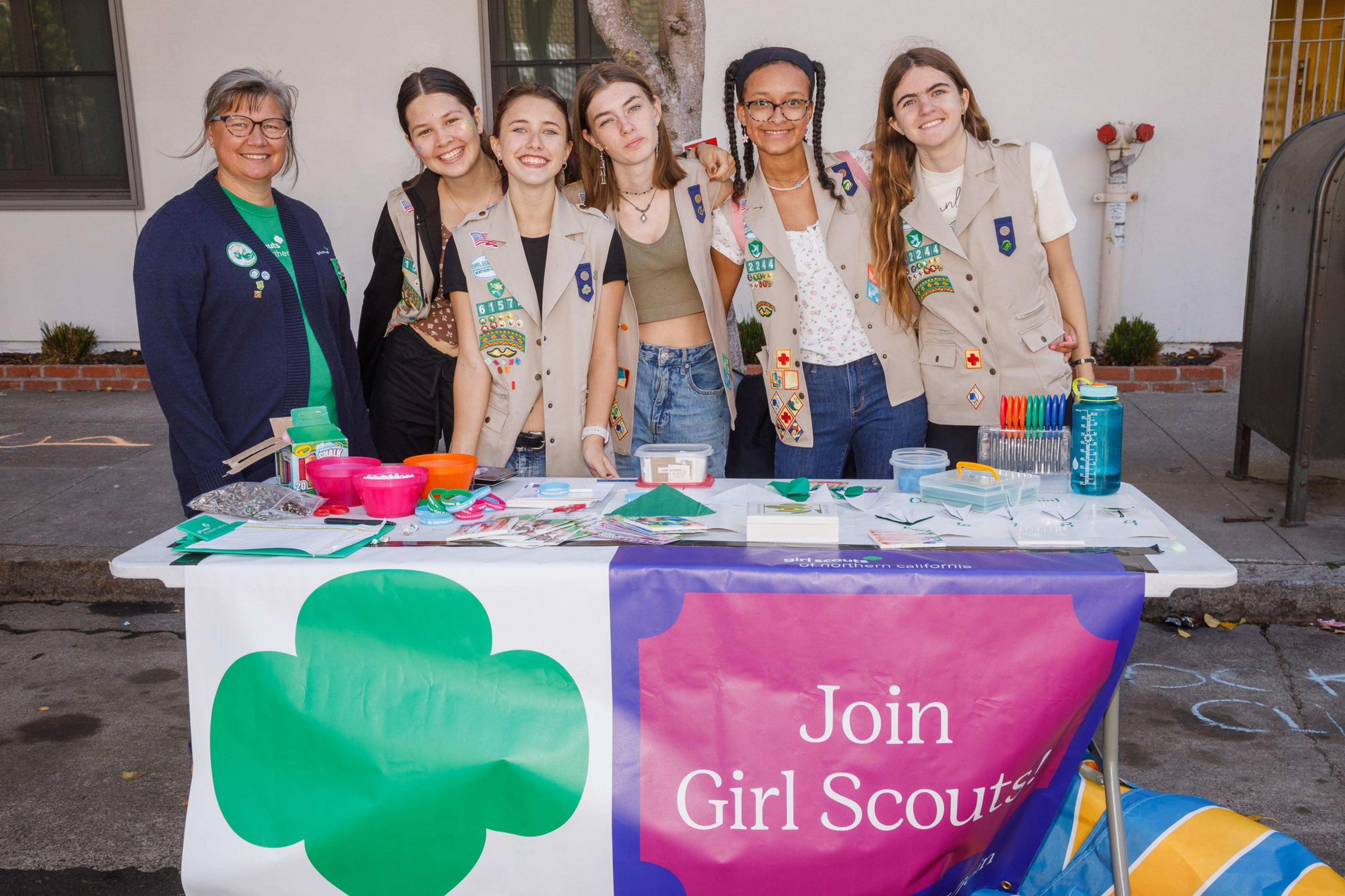 A group of Girl Scouts and a leader behind a table with promotional materials, smiling at a recruitment event.
