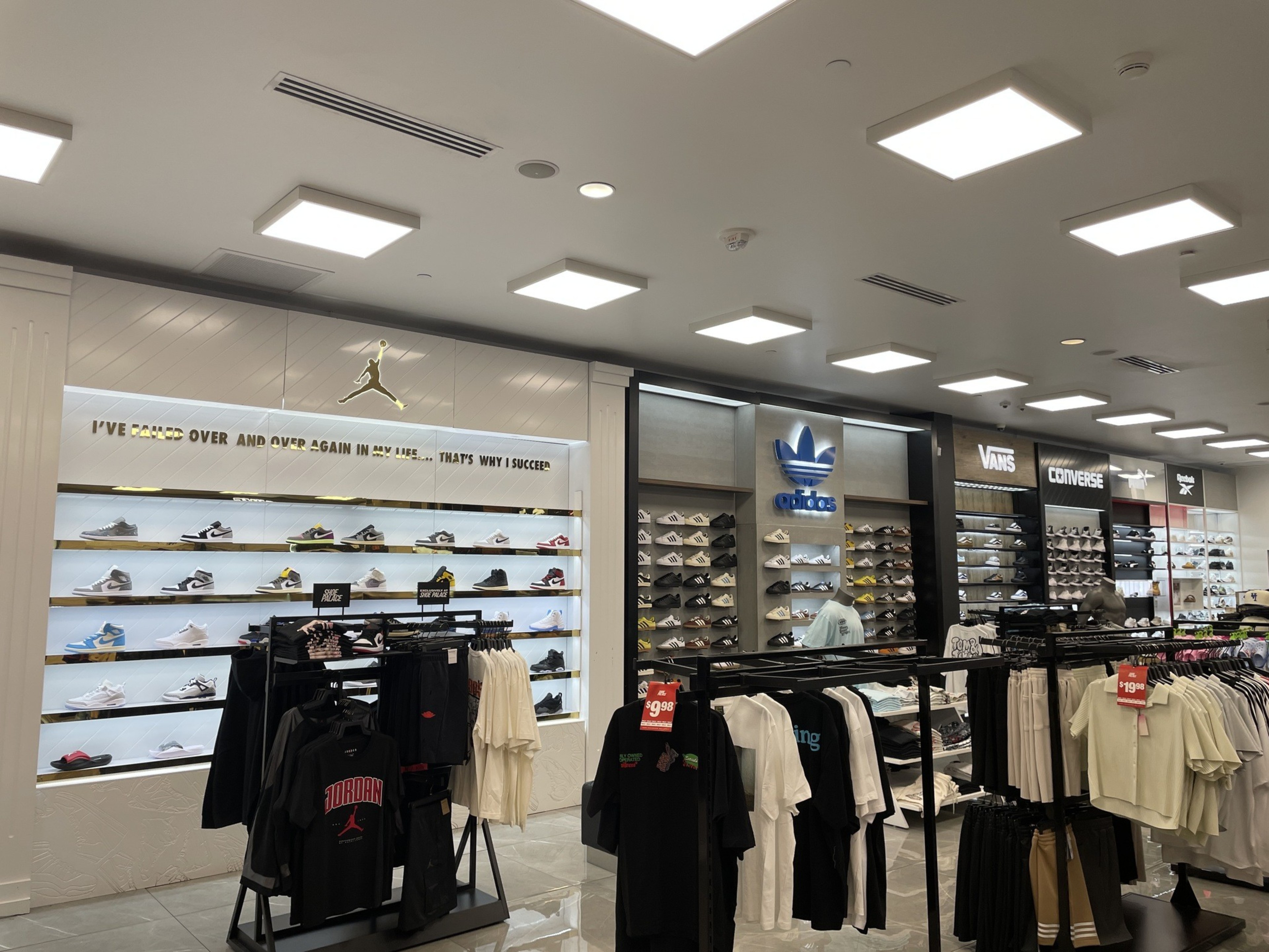 A brightly lit sneaker store displays shoes from brands like Jordan, Adidas, Vans, Converse, and Skechers with clothing racks in the foreground.