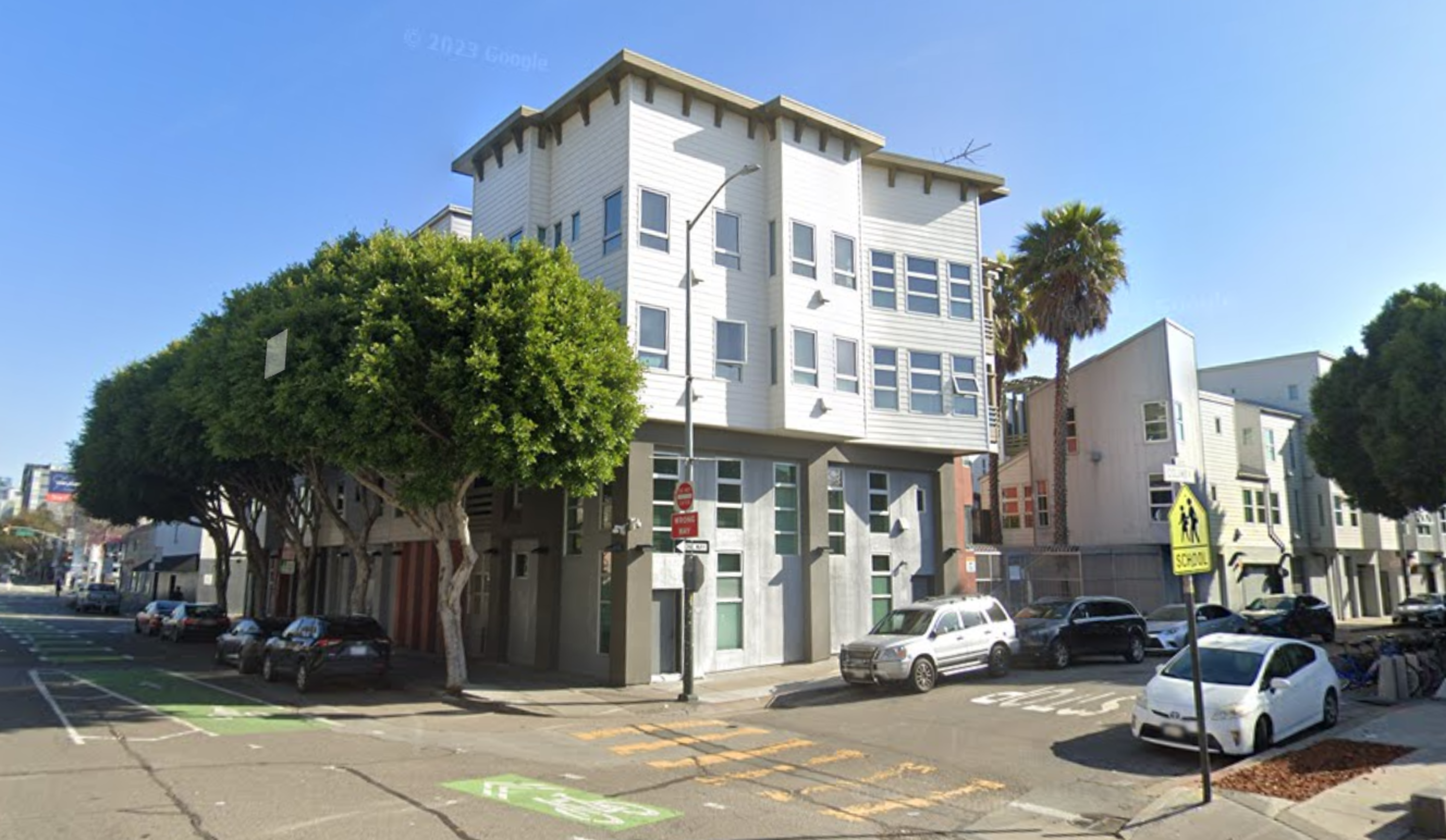 A modern four-story building stands at a street corner, surrounded by trees and parked cars under a clear blue sky.