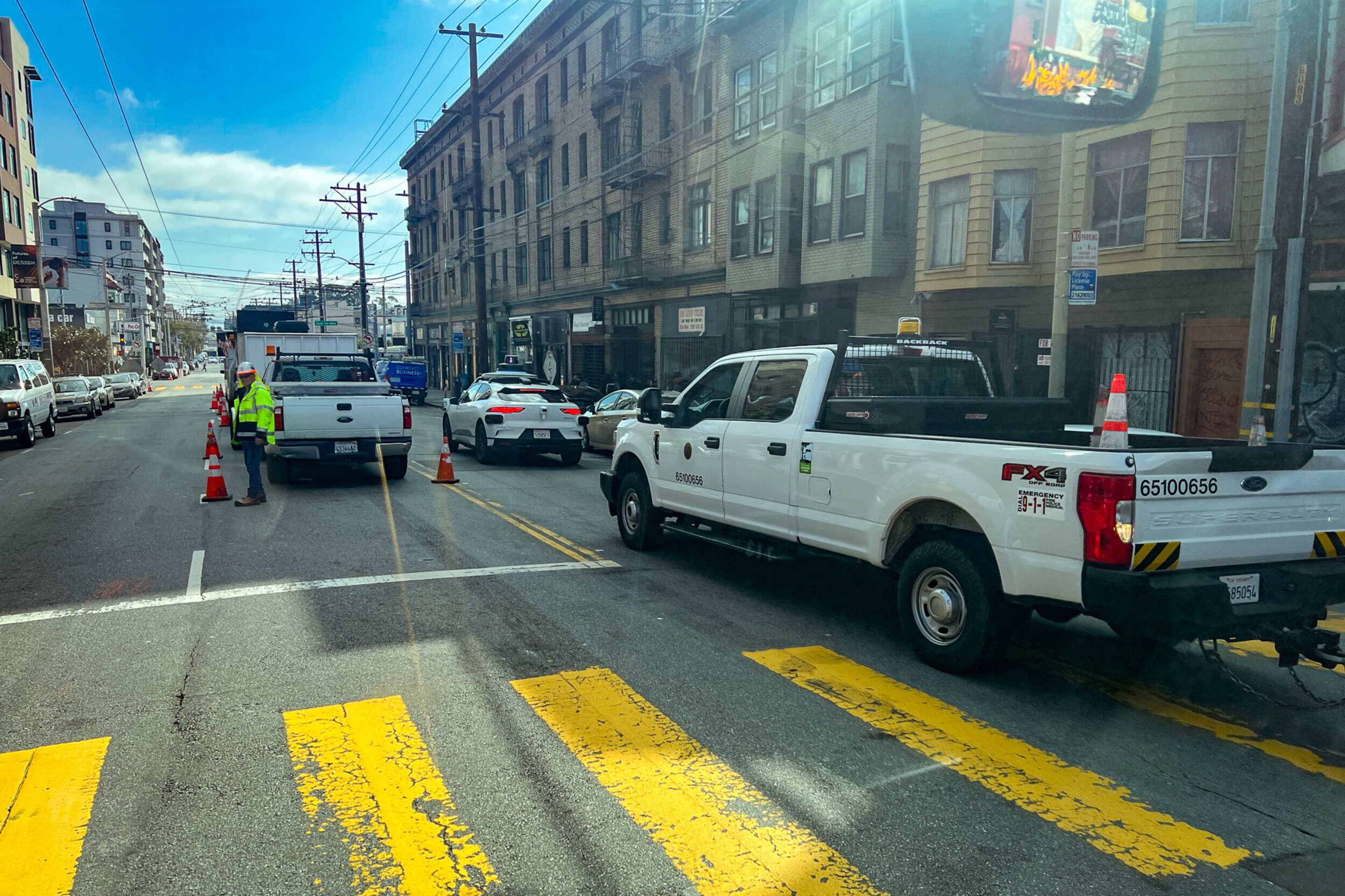 Two white utility trucks and a white sports car are parked on a city street with workers and orange traffic cones directing traffic.