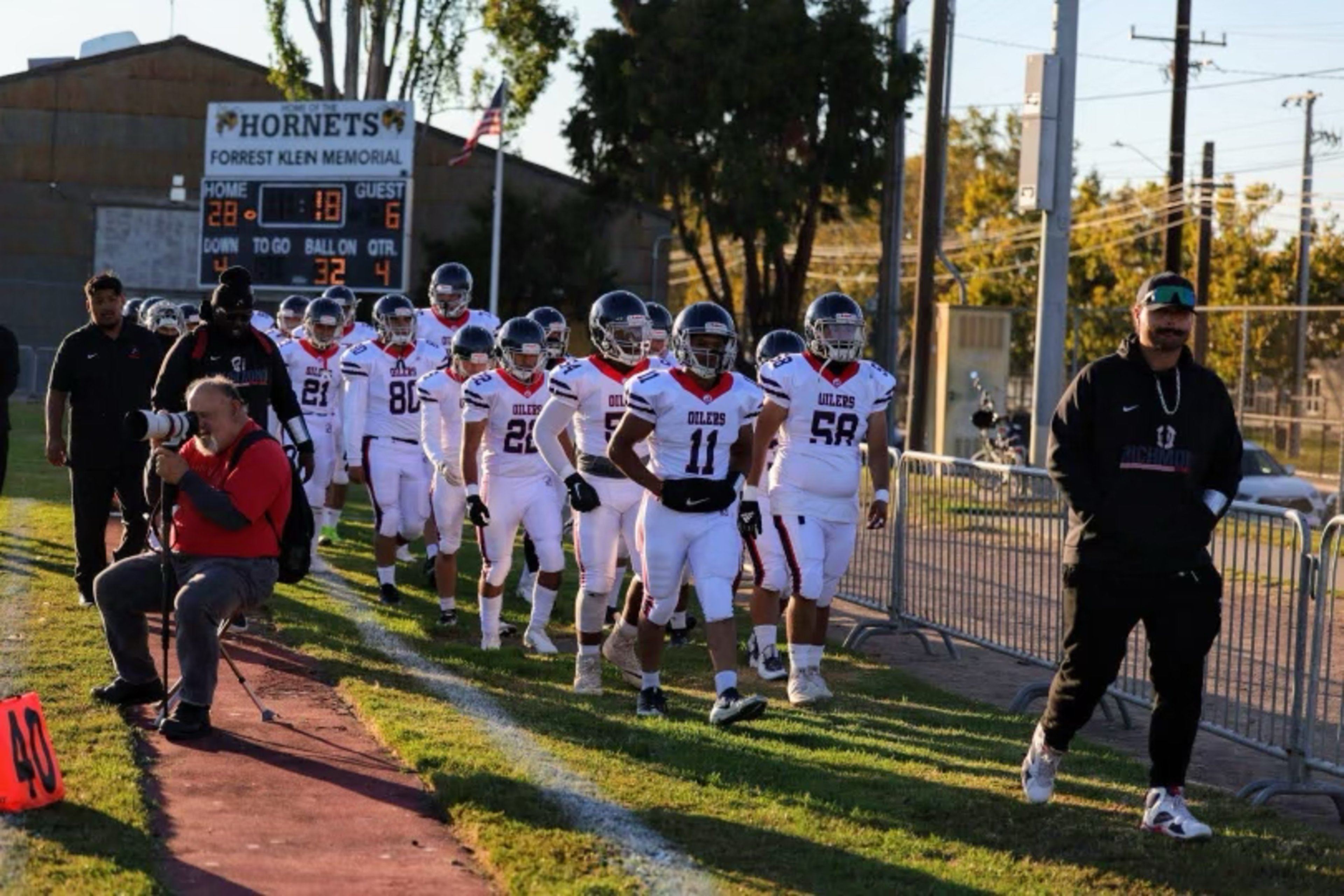 A football team in white uniforms walks on a field toward the camera, with a photographer sitting on the left and a scoreboard behind showing 28-6.