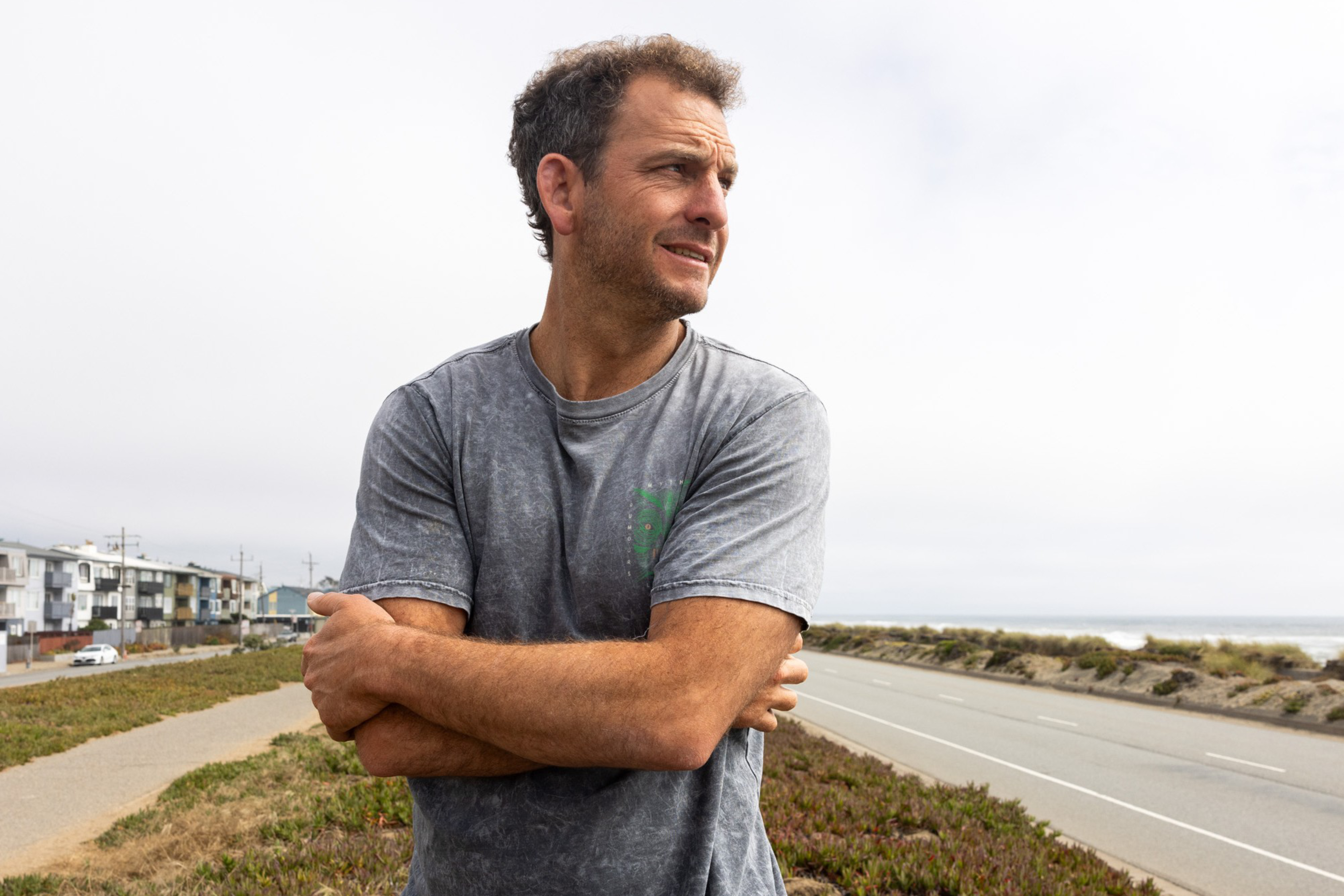 A man in a grey shirt stands with arms crossed, looking to his right. He is on a path near a road by the coast, with buildings and parked cars visible in the background.