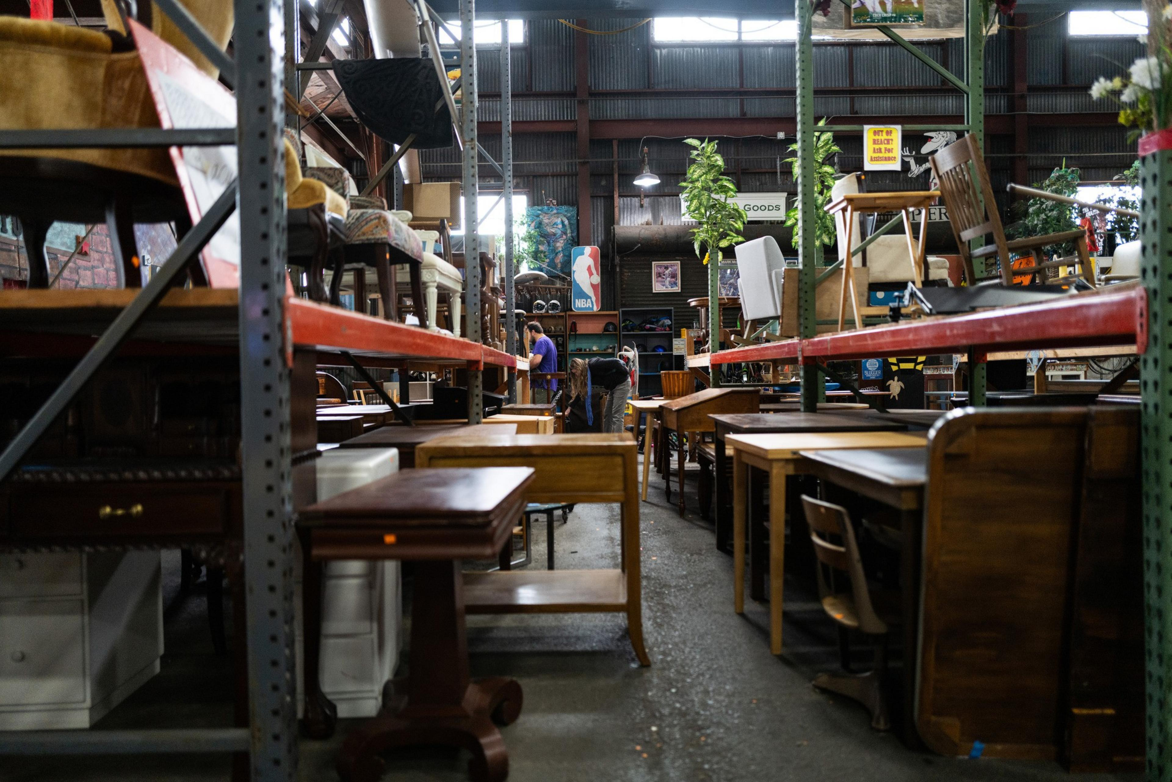 The image shows a warehouse filled with furniture, including chairs and tables on metal shelves. Two people are browsing, and a sign with the NBA logo is visible.