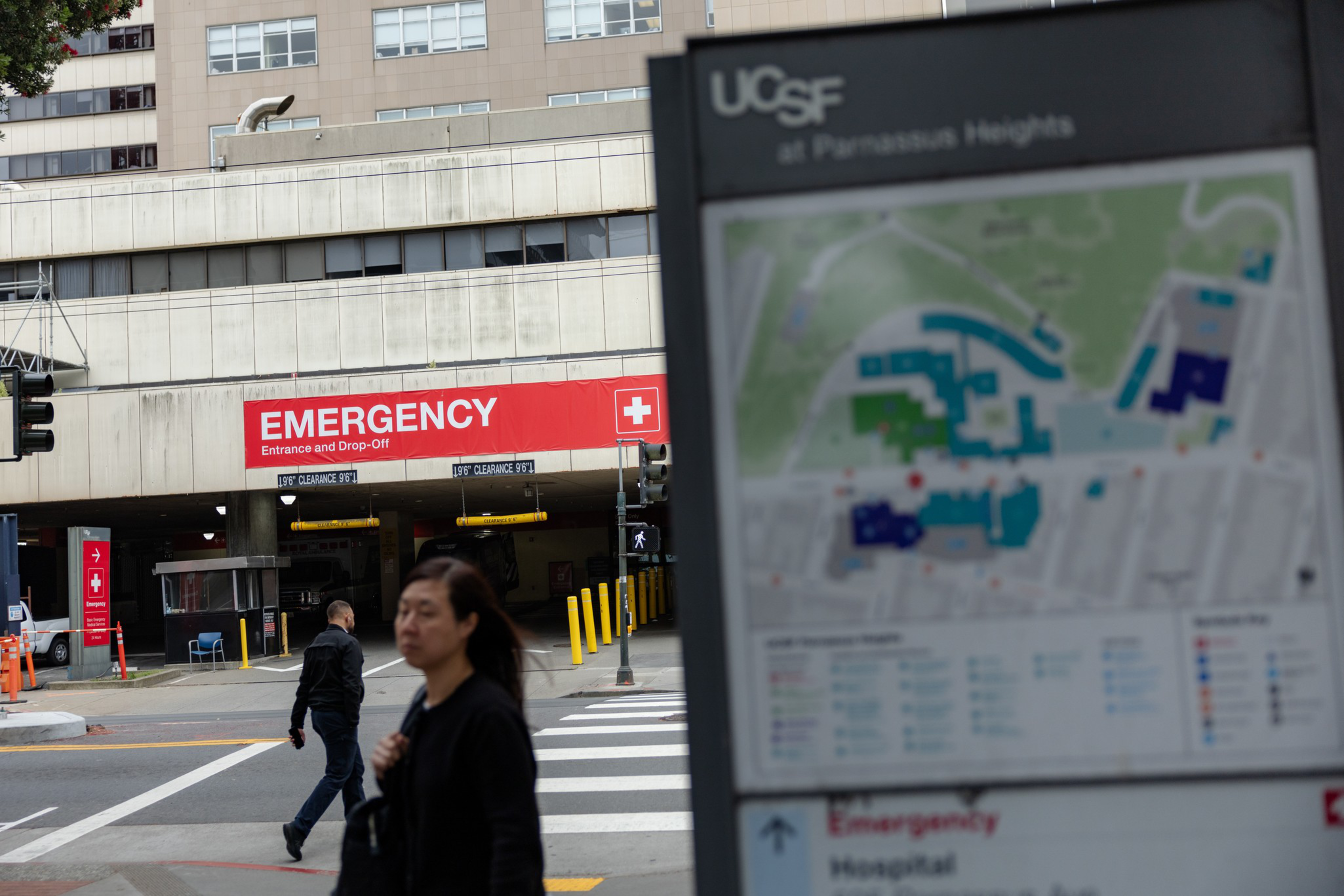 A hospital building with a red “EMERGENCY Entrance and Drop-Off” sign, a blurred campus map labeled UCSF in the foreground, and people walking nearby.