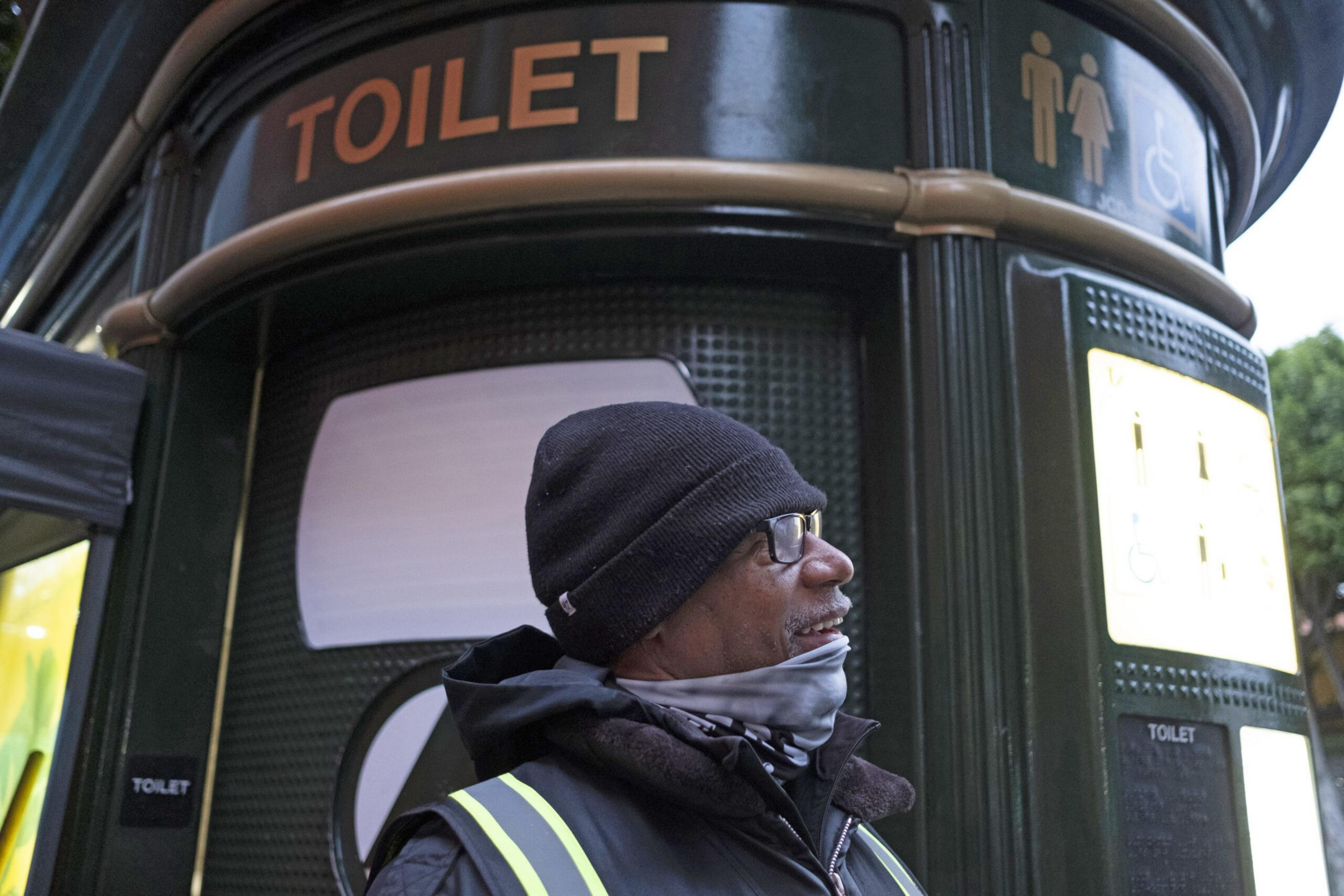 A man wearing a black beanie, glasses, and a dark jacket with reflective stripes stands near a public toilet with gender and wheelchair symbols.