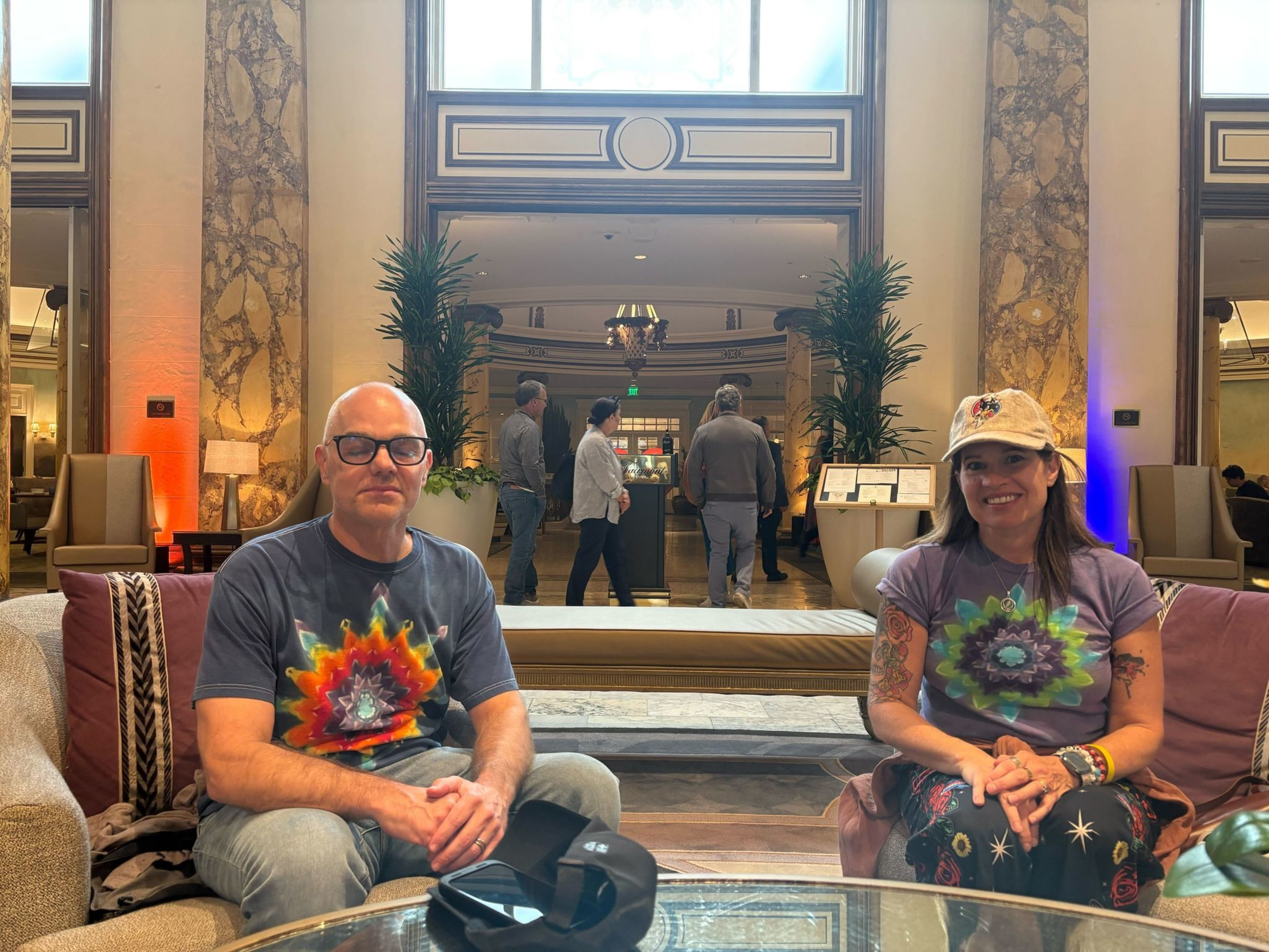 A man and a woman wearing colorful psychedelic t-shirts sit on a sofa in a grand lobby with marble columns and plants.