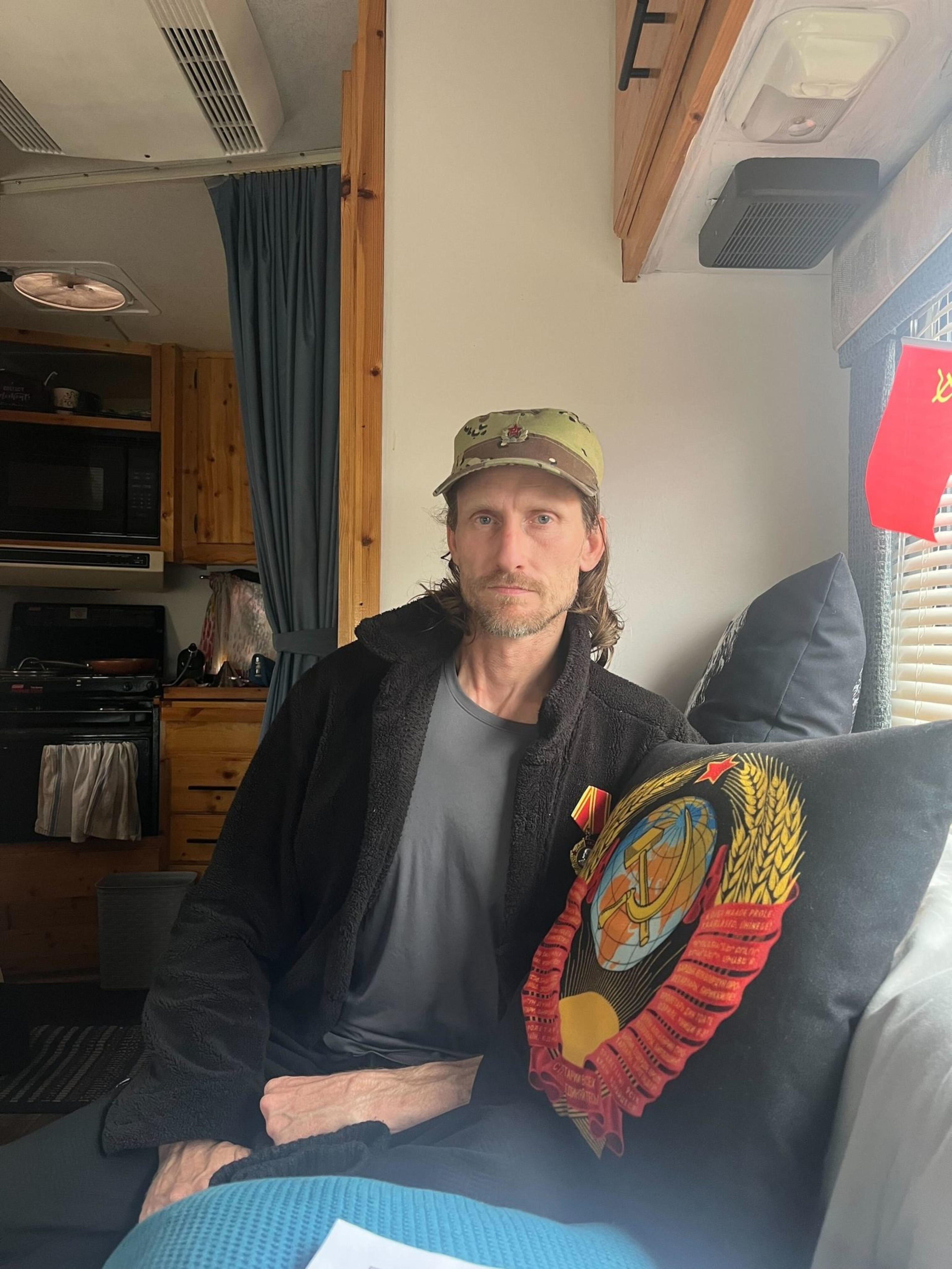 A person sits inside a wooden interior, wearing a hat and jacket. They are near a black pillow adorned with a colorful emblem and next to red fabric.